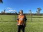 Tracie Smith’s father standing in a sunny field wearing an orange and black jersey, smiling with a backdrop of green grass, trees, and a clear blue sky.