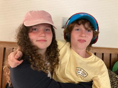 A candid photo of Halen wearing headphones and a blue cap, sitting next to his sister Addison, who is wearing a pink cap, as they relax together on a bench.