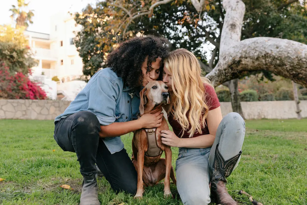 Two women kissing a dog in a green field