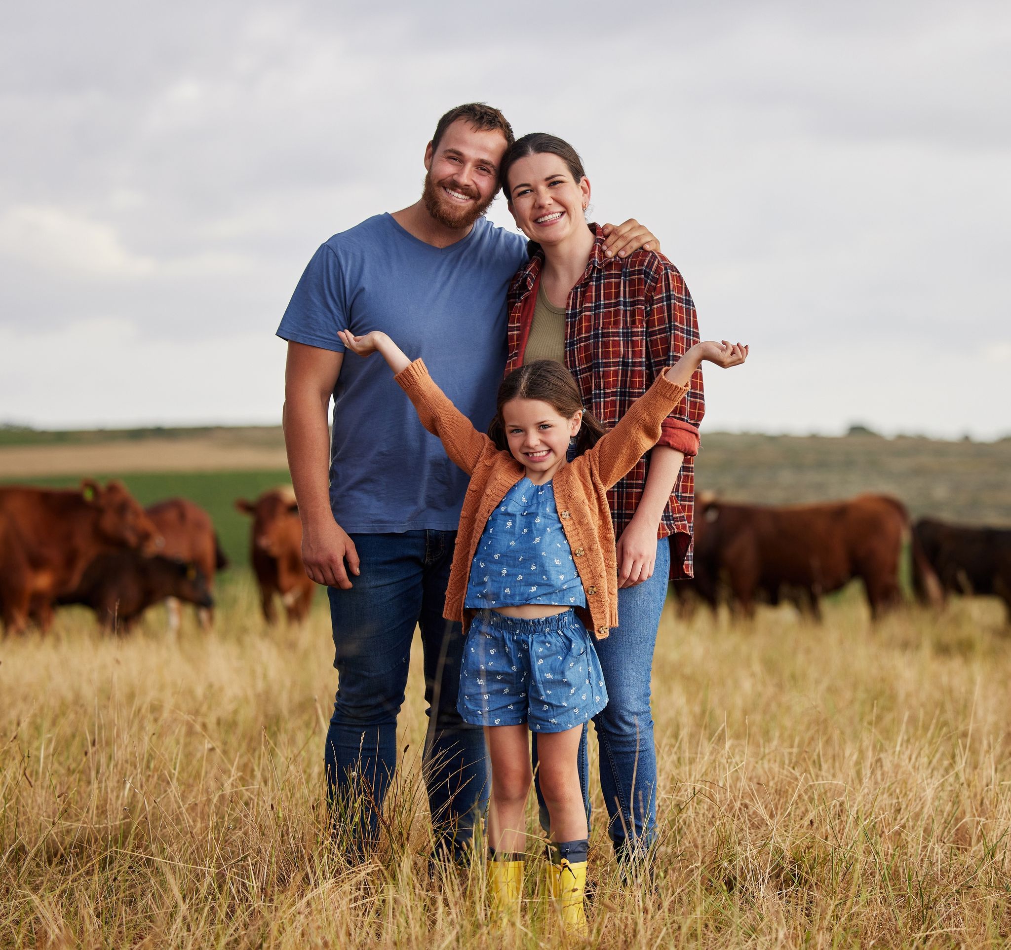 A couple with a young daughter standing in a dry grass field with cows behind them