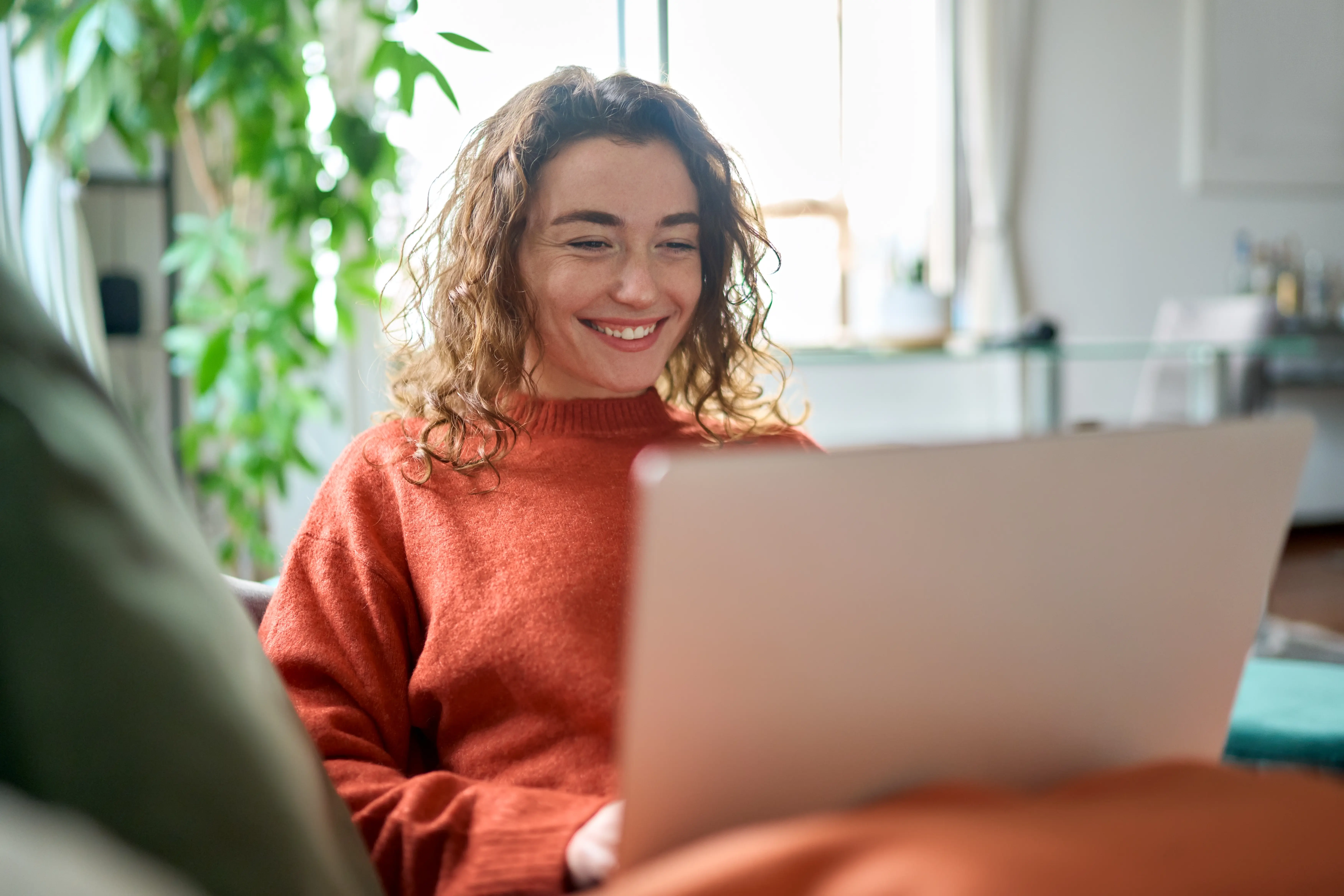 Young woman in orange sweater checking her laptop at home on a bright day