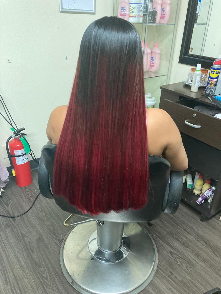 A woman is seated in a salon chair with her back facing the camera. She has long, straight hair that transitions from one color at the roots to a darker color at the ends. The hair is smooth and shiny, cascading down her back, and is trimmed evenly at the bottom.