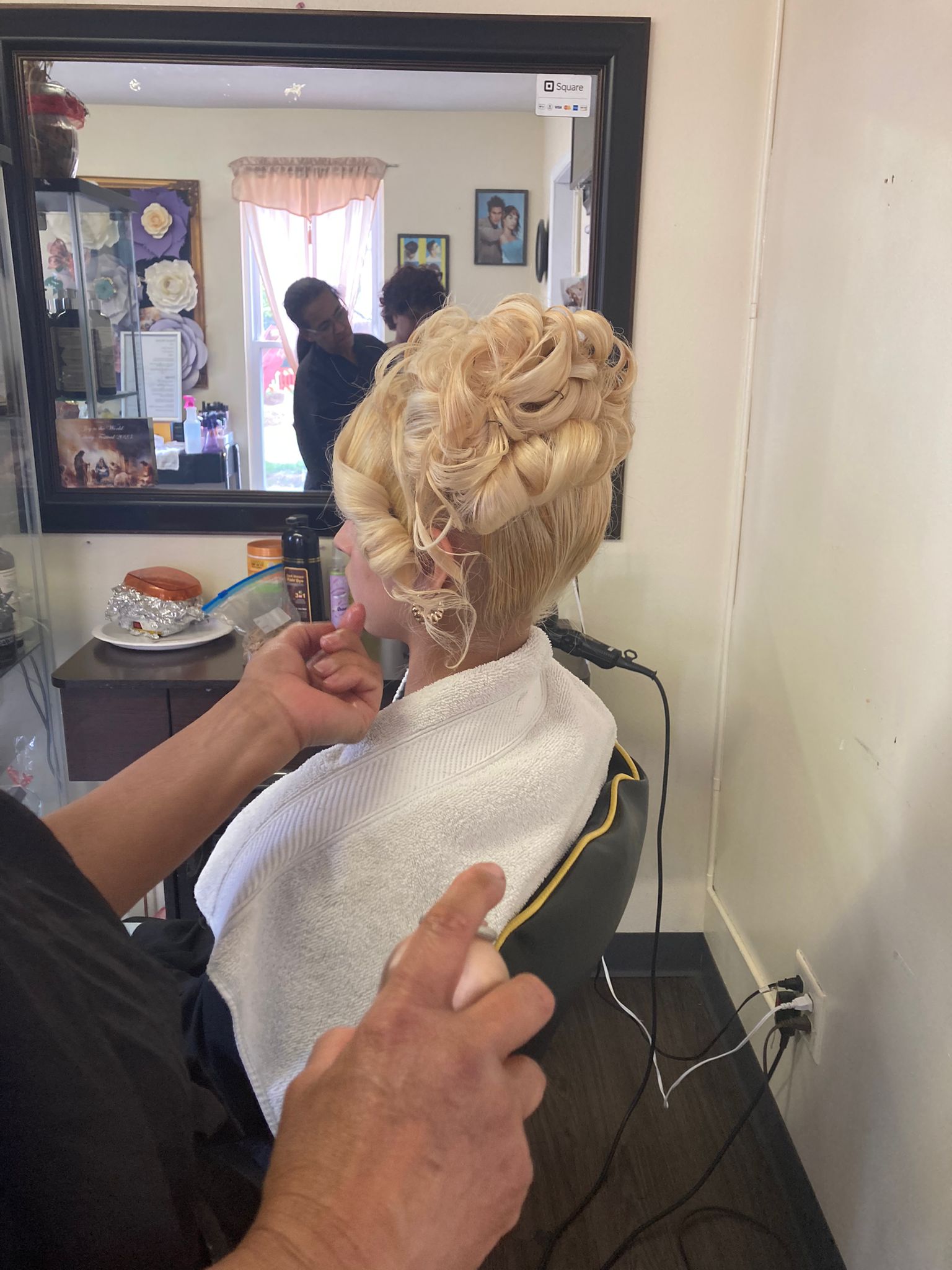 A woman with a sophisticated updo featuring tightly coiled curls meticulously arranged at the back of the head. The blonde hair is styled into voluminous, overlapping loops that create a detailed, elegant pattern. A loose, curled strand falls gracefully near the ear, adding a touch of softness to the overall look.