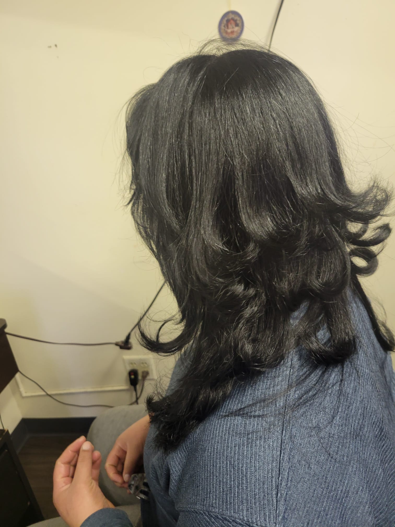 A side view of a person with long, layered hair styled with outward feathered ends, creating a voluminous and flowing look. The hair shines with a healthy gloss, and the layers give a textured, dynamic appearance.