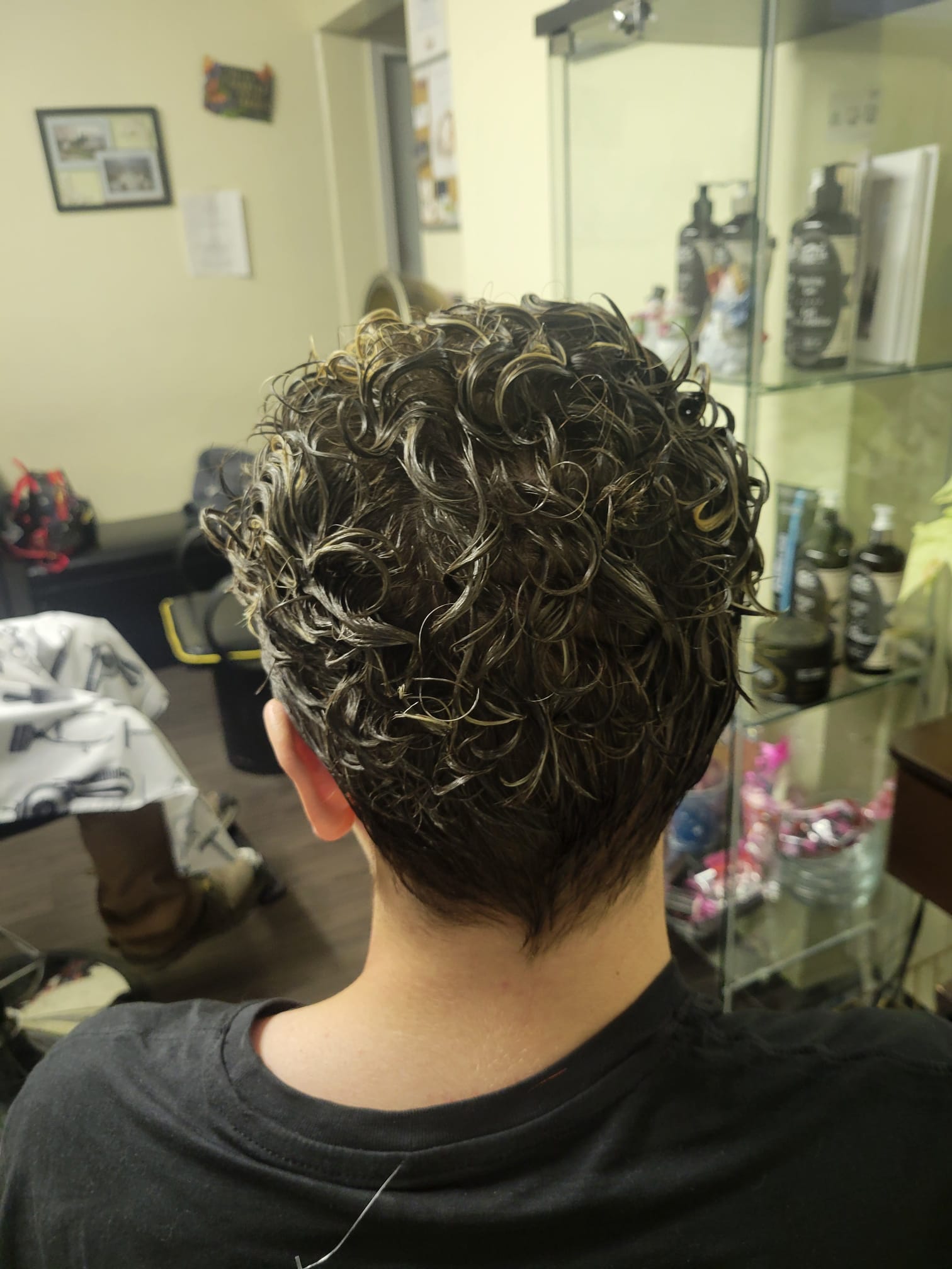 short, hair styled into tight, glossy curls created through a perm. The back view showcases neatly defined curls with a slightly tapered cut near the neckline.