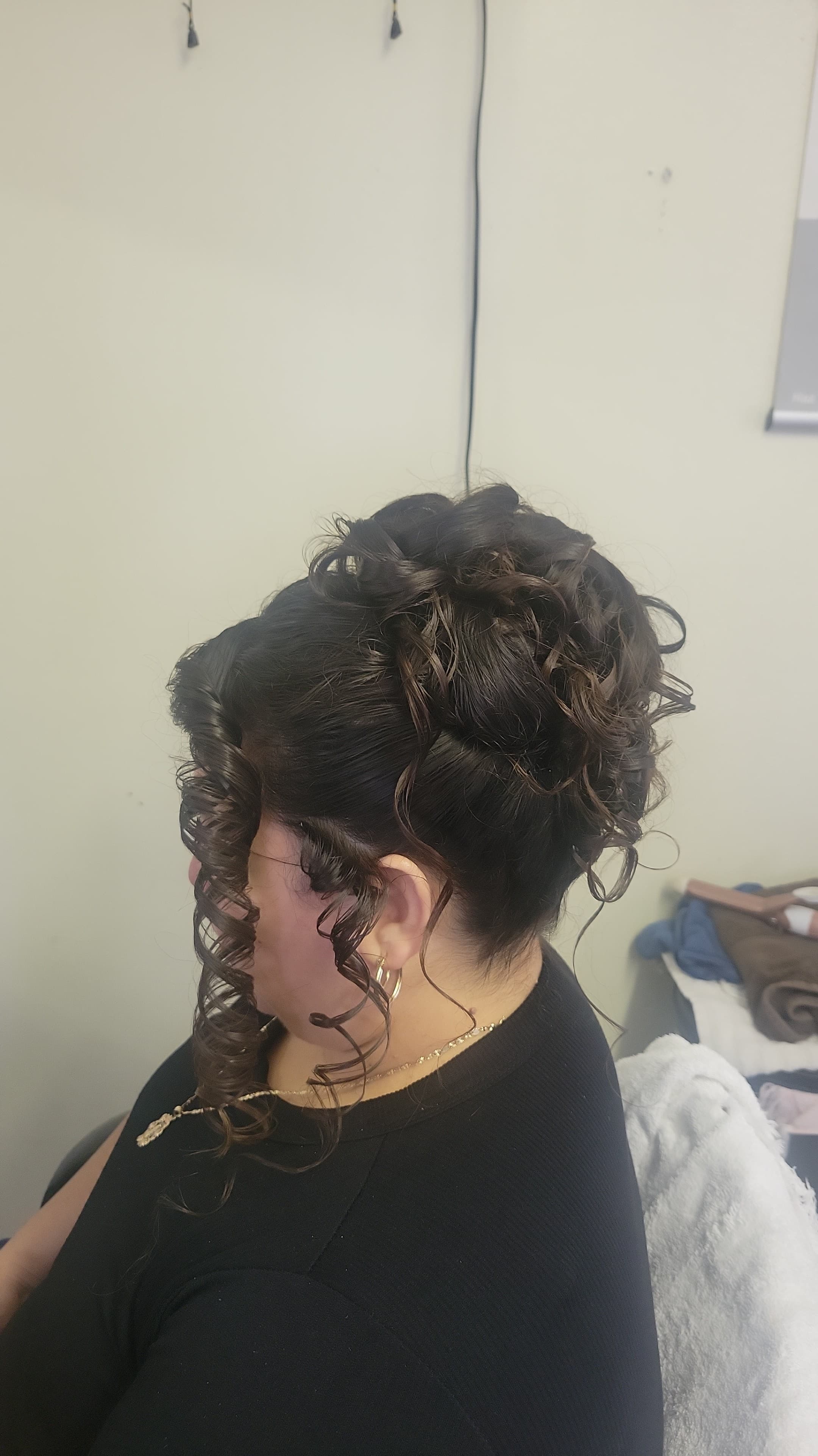 The hair is gathered high on the back of the head into a voluminous cluster of pinned curls. A long, tightly defined ringlet curl cascades down the side of the face, with smaller, delicate curled tendrils framing the ear.