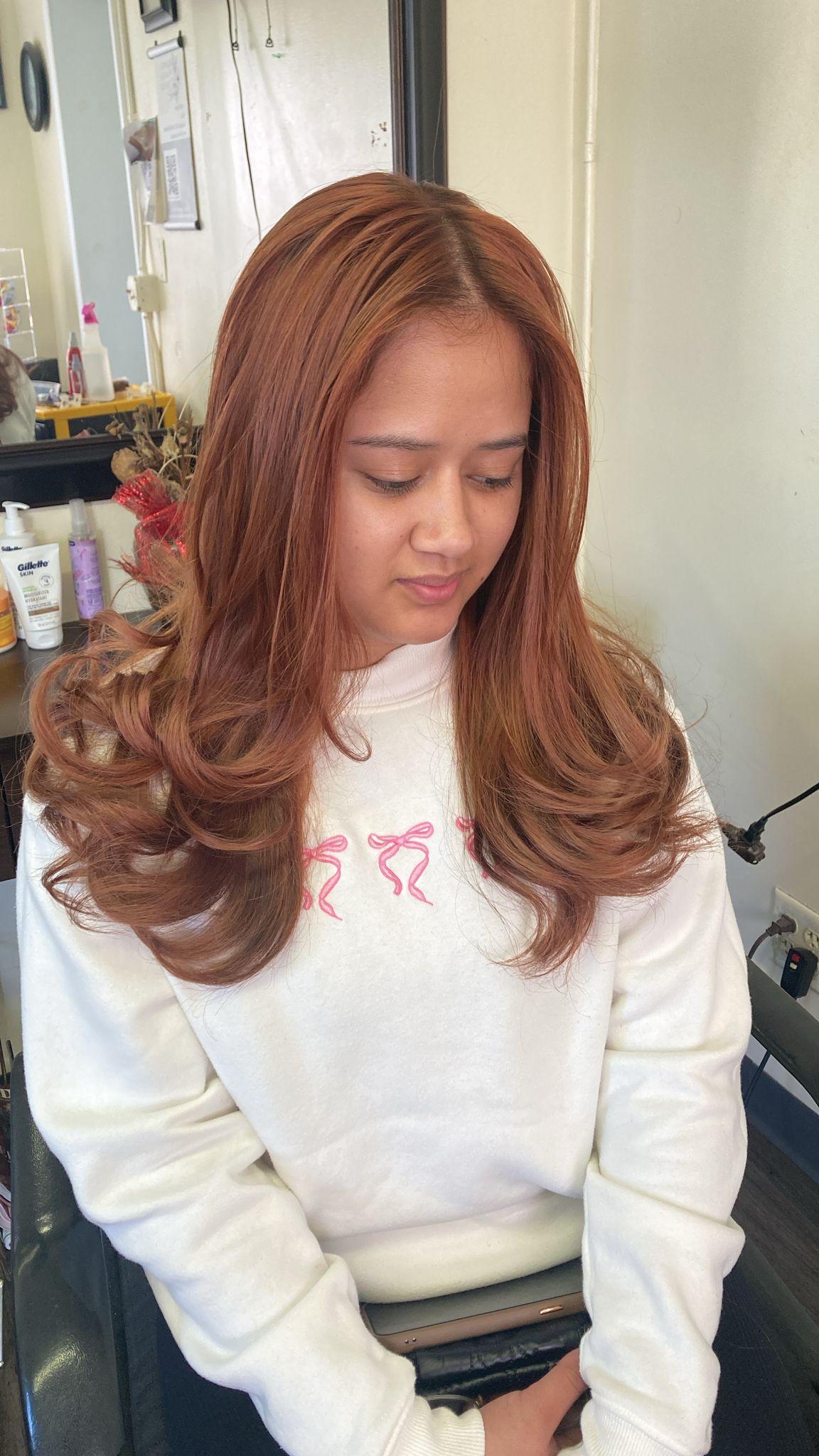 A woman with copper-toned hair sits in a salon chair, looking down. Her hair is styled in a smooth blowout with large, soft curls at the ends, creating a full, voluminous look. The hair is parted in the center.