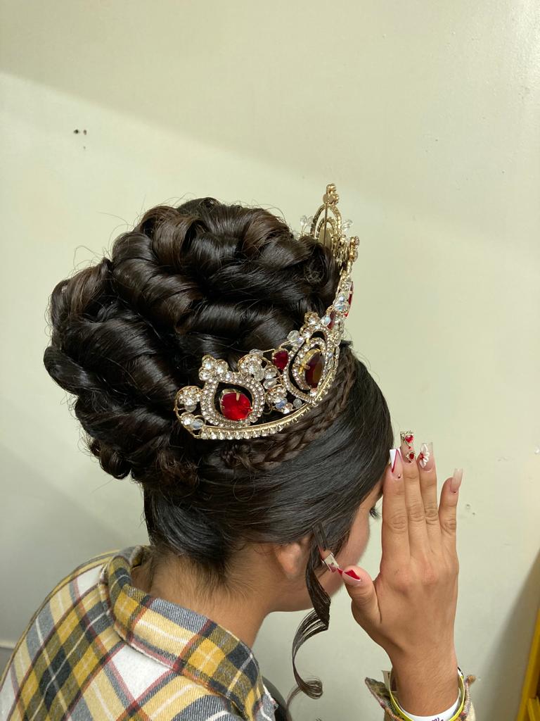 Woman with an intricate hairstyle featuring tightly coiled curls arranged into a voluminous updo. The curls are meticulously styled and pinned up, creating a high, textured look. A single braid wraps around a decorative tiara with gemstones is placed on top, adding a regal touch. The person wearing the hairstyle has their left hand raised to their face, partially covering it.