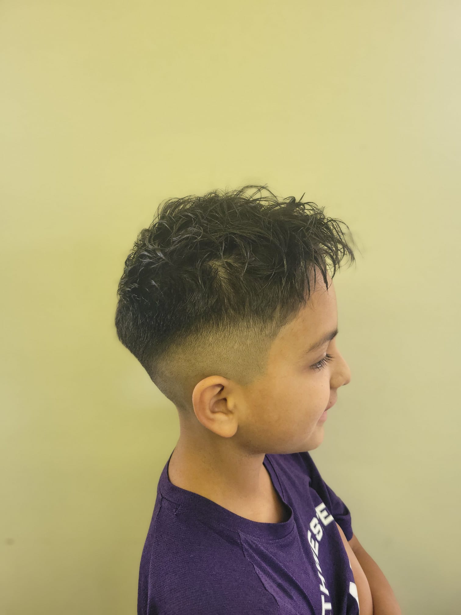 A young boy with slightly messy textured hair styled on top, featuring a sharp low fade on the sides and back. The haircut transitions smoothly from the skin fade to the longer hair on top.