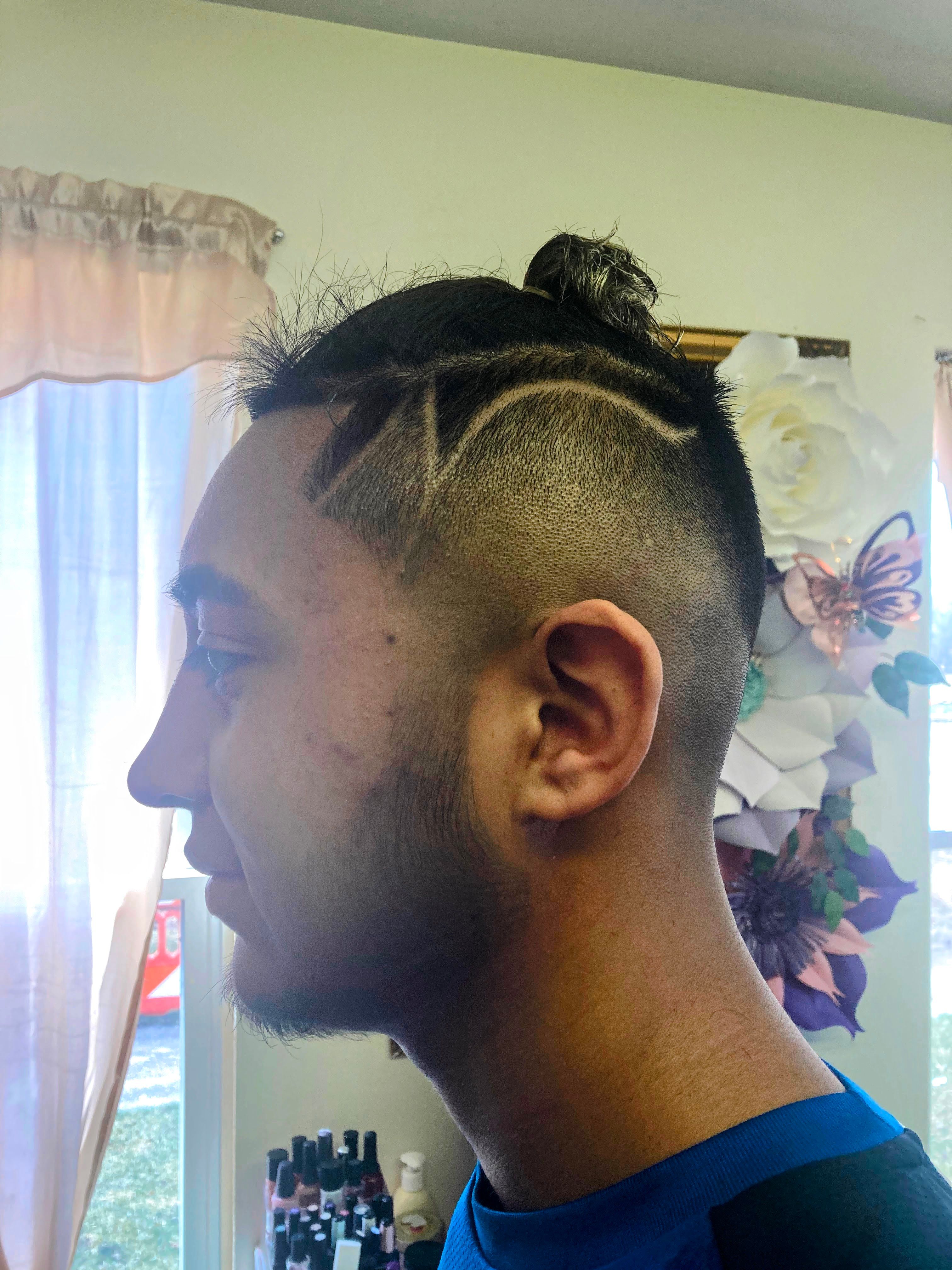 A man with a unique haircut. The sides of the head are closely shaved, with a high fade leading to longer hair on top, which is tied into a small bun. The shaved sides feature an intricate design of geometric lines and curves