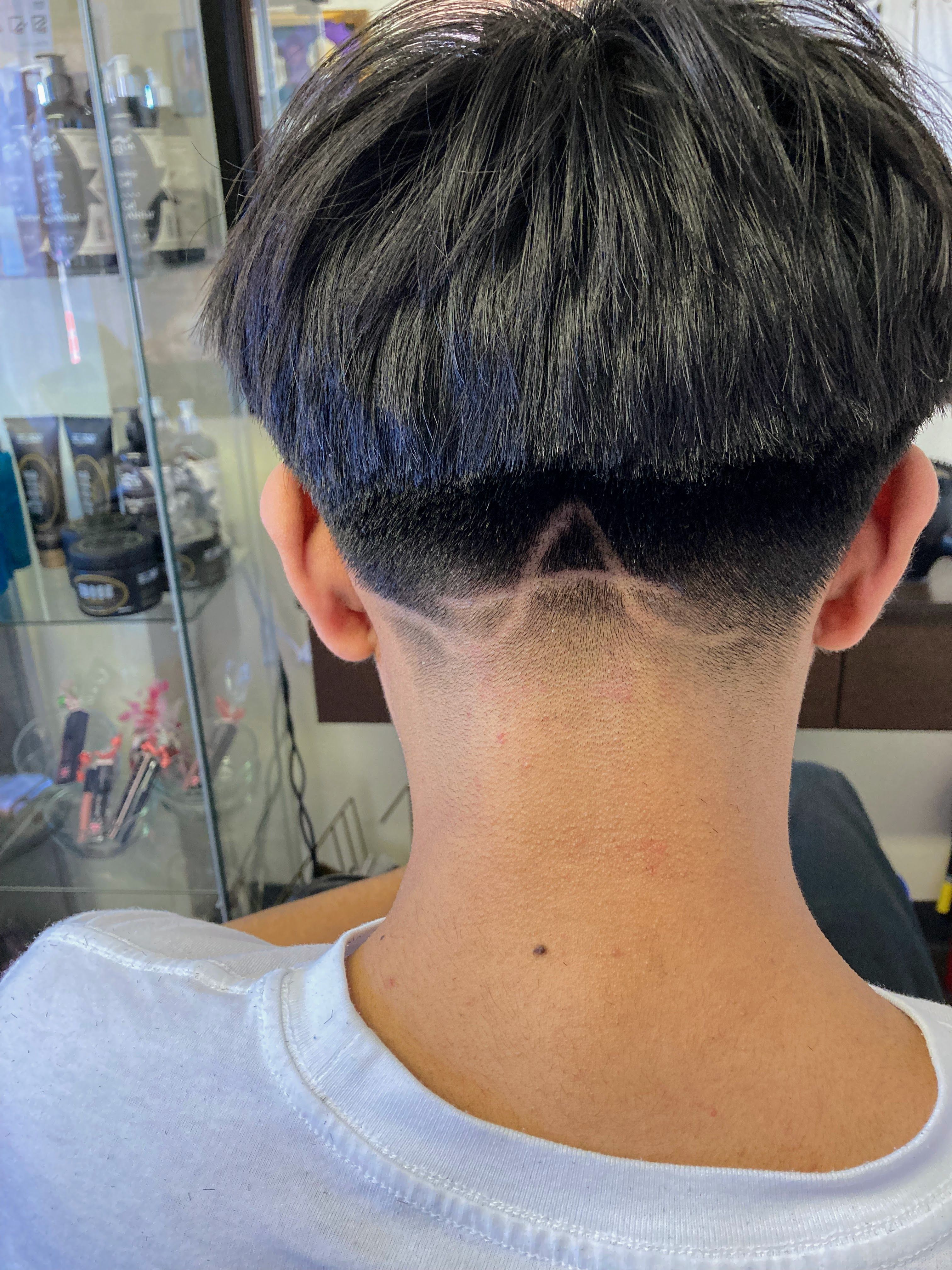 Back of a person’s head with a bowl cut hairstyle. The hair is thick and straight, with the longer top section ending in a blunt line above the ears. The undercut is closely shaved, featuring a precise, symmetrical geometric design in the shape of a triangle at the nape of the neck.