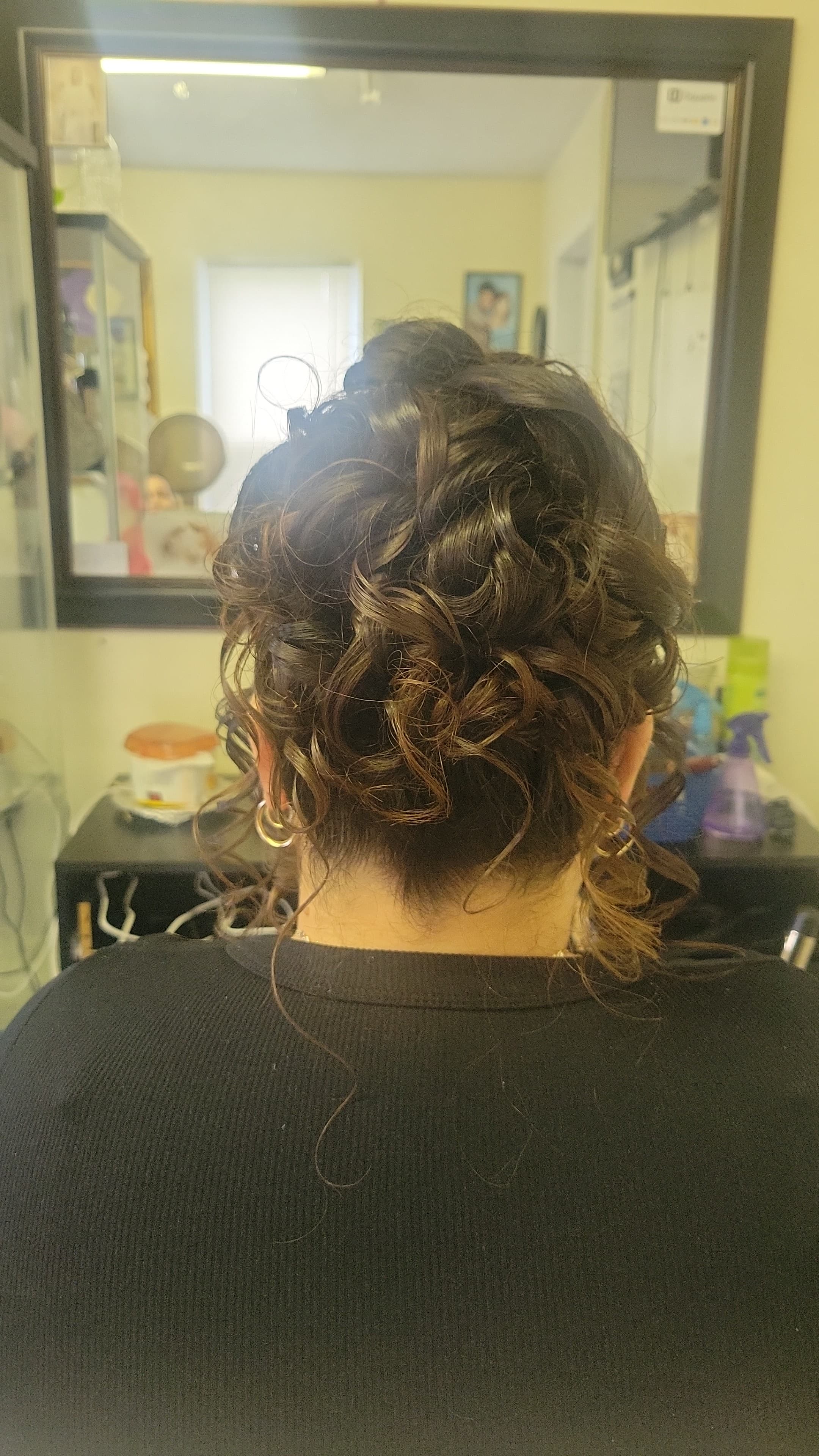 The dark brown hair is cleanly swept up away from the nape of the neck, forming a rounded, highly textured bundle of pinned ringlets. A few wispy, single-stranded curly tendrils fall loosely at the base of the neck.