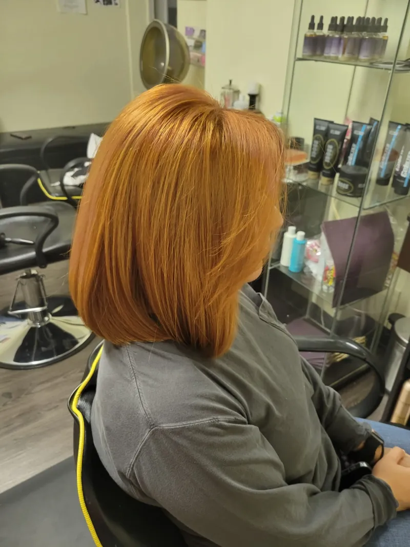 Mujer sentada en una peluquería, vista de perfil. Tiene el pelo liso hasta los hombros, teñido de un cálido color naranja cobrizo. Lleva una camiseta de manga larga.