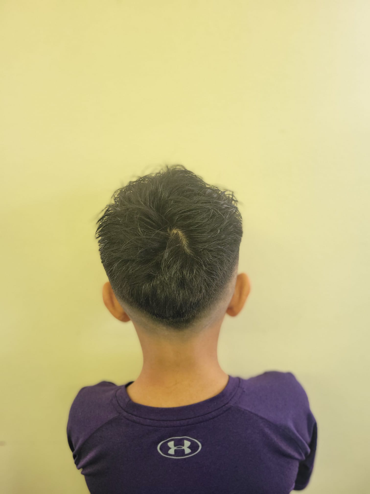 A young boy is shown from the back. The hairstyle features a low skin fade that tapers smoothly from the base of the neck up to the crown, where the hair transitions into a textured, slightly messy crop. The hair on top is longer, adding volume.