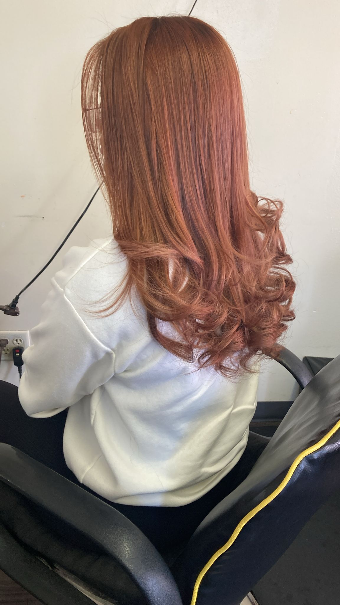 A woman is seated in a salon chair with her back to the camera. Her hair is a warm copper color, styled in smooth, voluminous curls that begin around mid-length and cascade down to the ends. The top of her hair is straight and sleek, creating a contrast with the bouncy curls at the bottom.