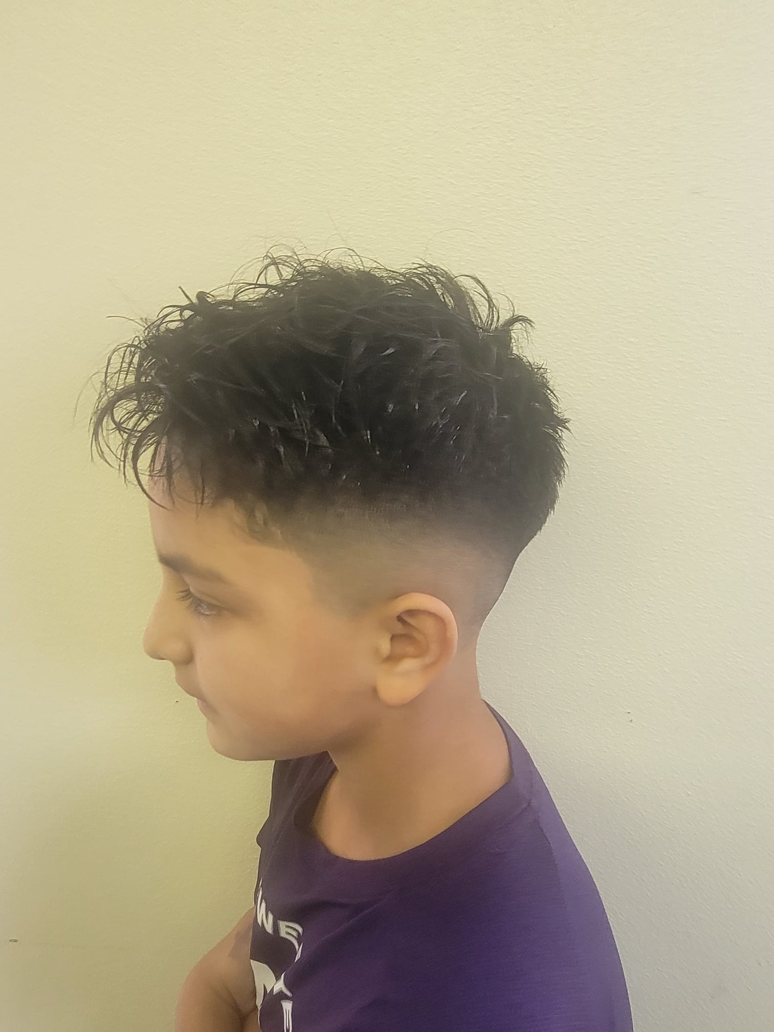A young boy with slightly messy textured hair styled on top, featuring a sharp low fade on the sides and back. The haircut transitions smoothly from the skin fade to the longer hair on top.