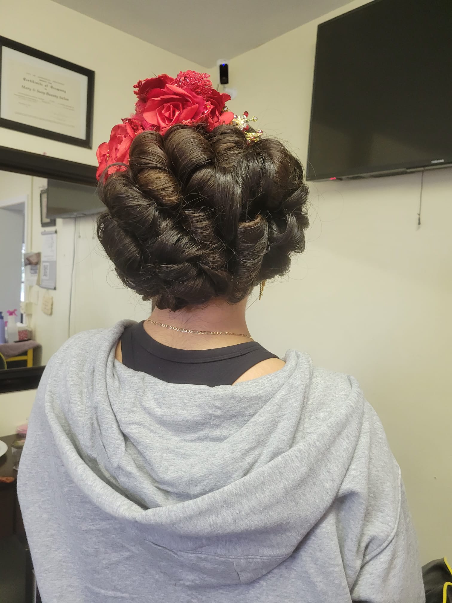 A woman with dark hair styled in a low braided bun adorned with large, vibrant silk flowers. The flowers are intricately detailed, with a mix of petals, lace, and embellishments.