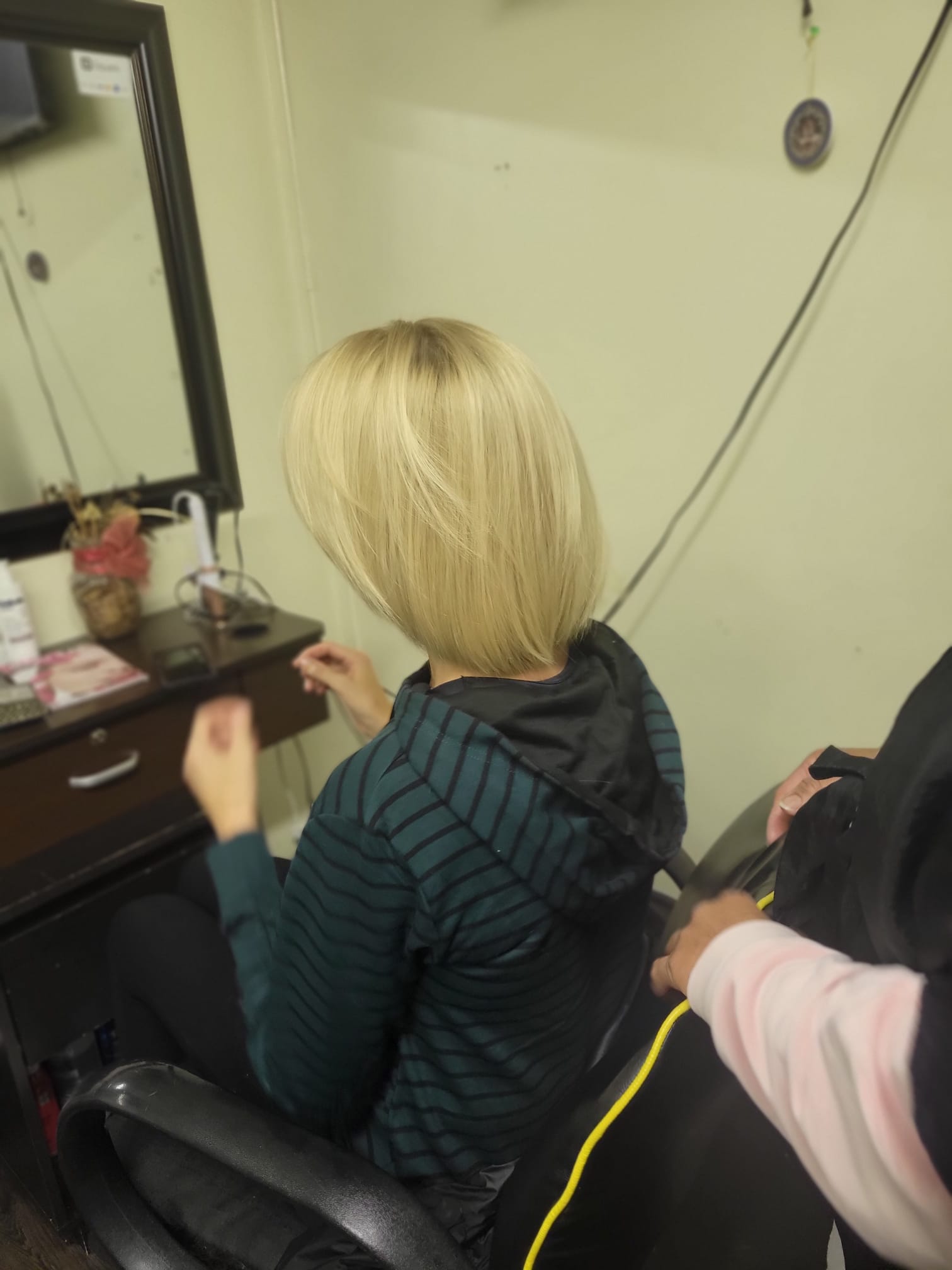 Straight, sleek, shoulder-length layered bob. The cut has a slightly rounded shape at the back, adding volume, with a smooth and polished texture. She is seated in a salon chair, with a stylist behind her.