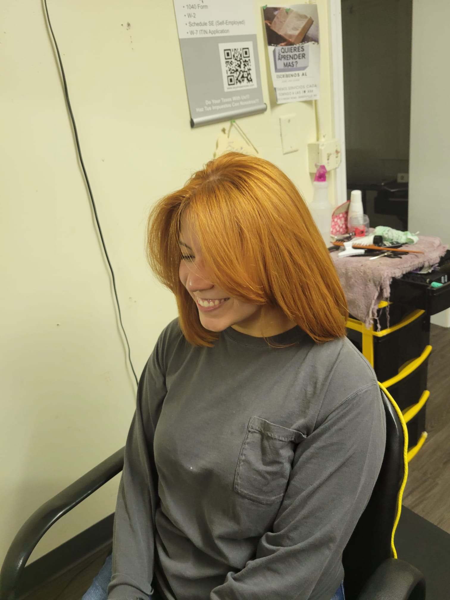 A person sitting in a hair salon, viewed from the front and slightly to the side. They have straight, shoulder-length. They are smiling and wearing a long-sleeved shirt with a pocket on the chest.