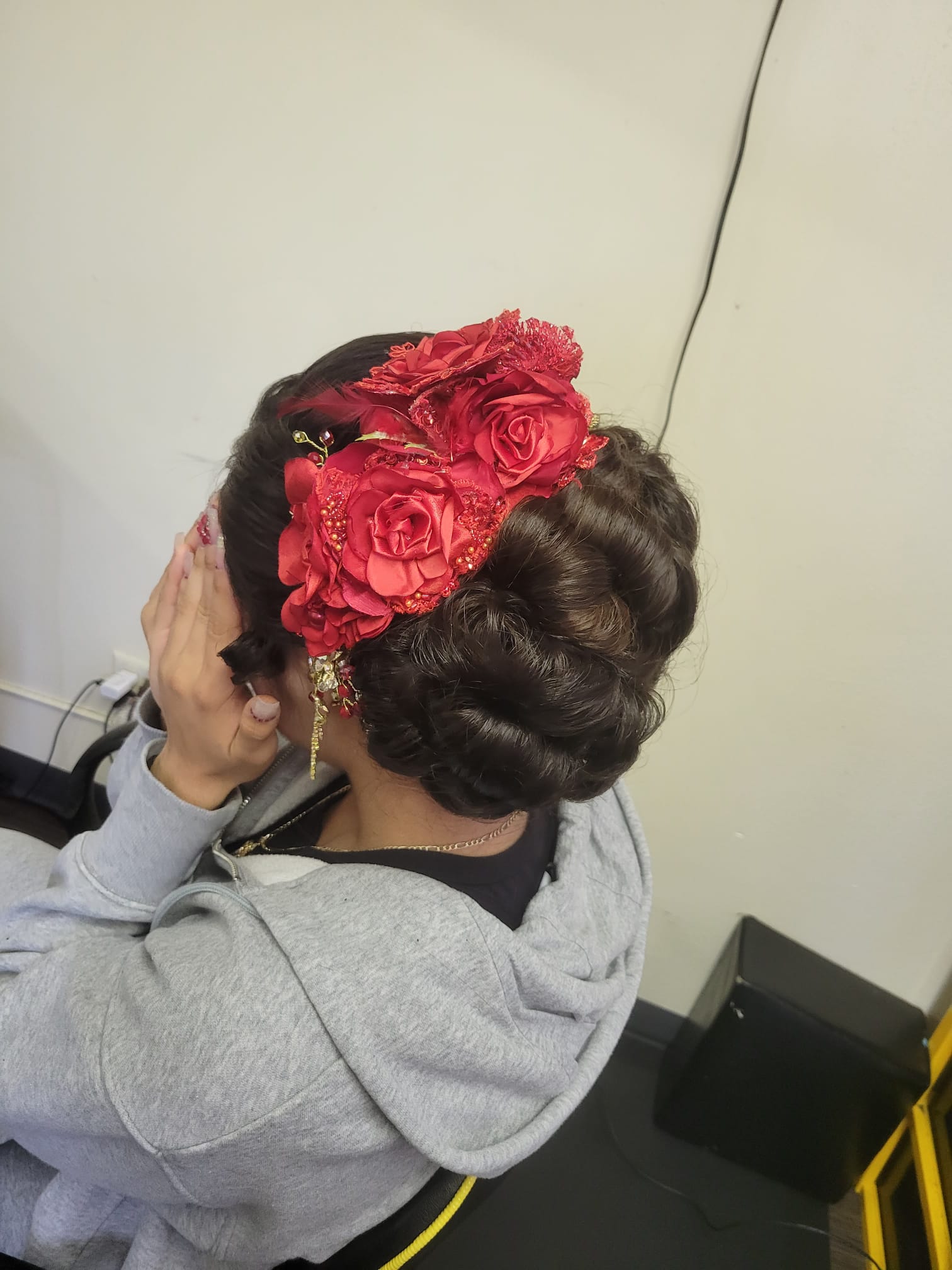 A woman with dark hair styled in a low braided bun adorned with large, vibrant silk flowers. The flowers are intricately detailed, with a mix of petals, lace, and embellishments.