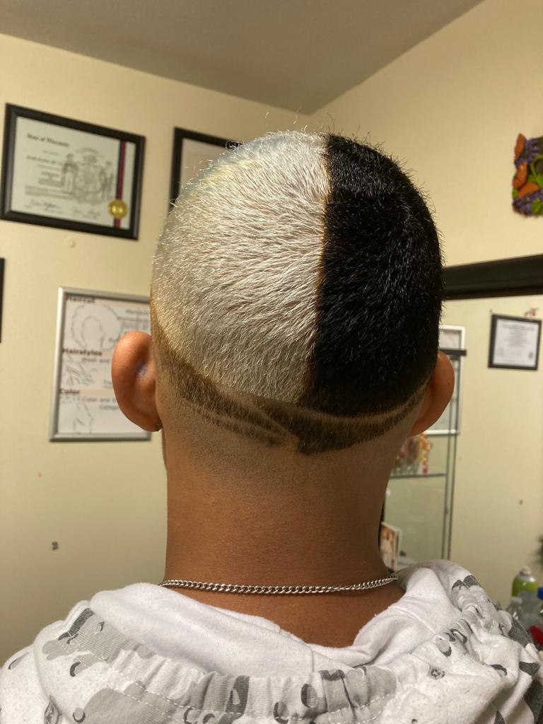 Back view of a person’s head. The hair is split vertically down the middle, with the left side bleached white and the right side left black. At the bottom of the head, there is a precise and intricate shaved design featuring a curved line and a small triangular shape