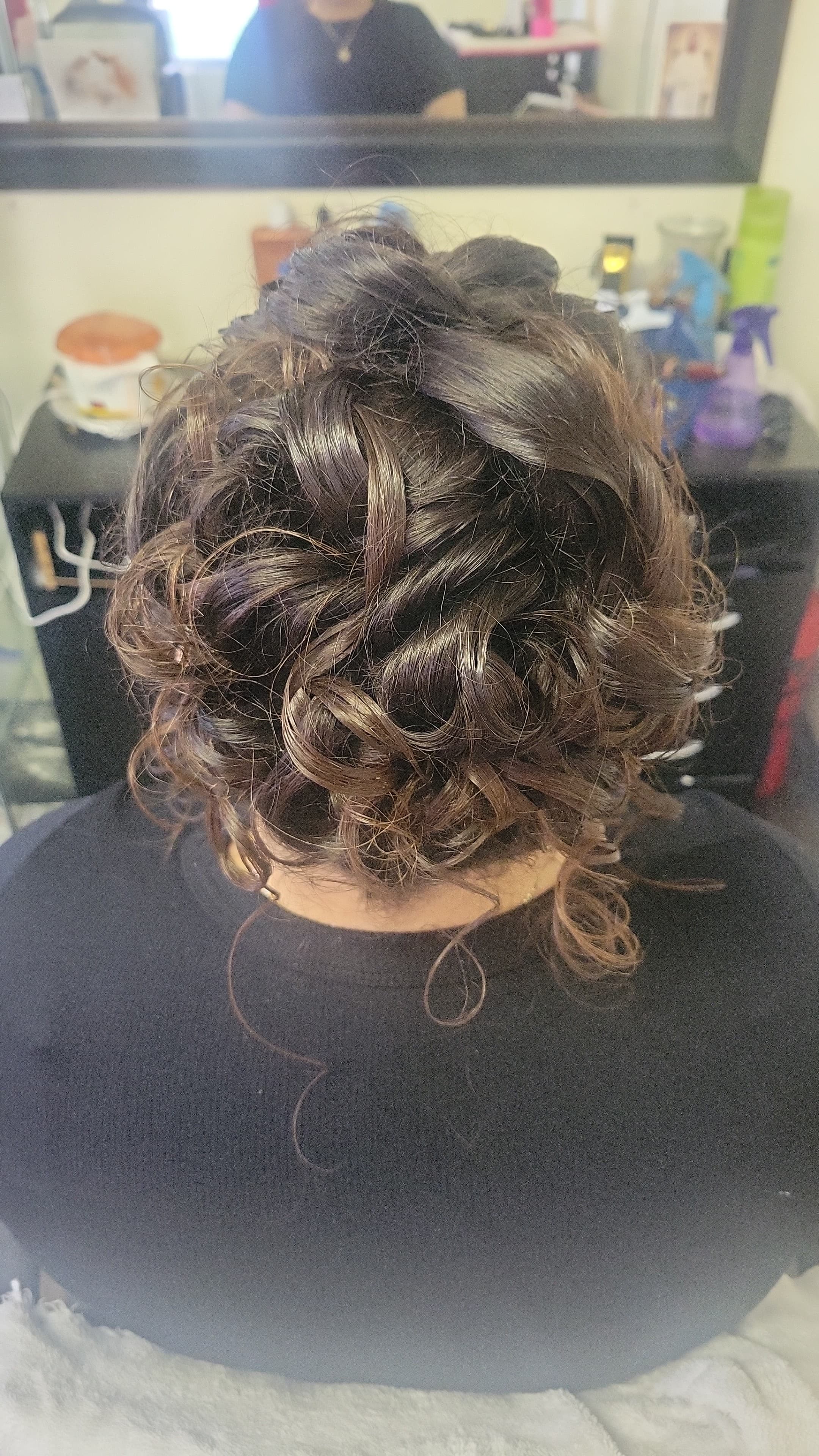 The dark brown, glossy hair is intricately arranged and pinned into an elegant arrangement of overlapping loops, swirls, and tight curls that create height and volume across the crown.