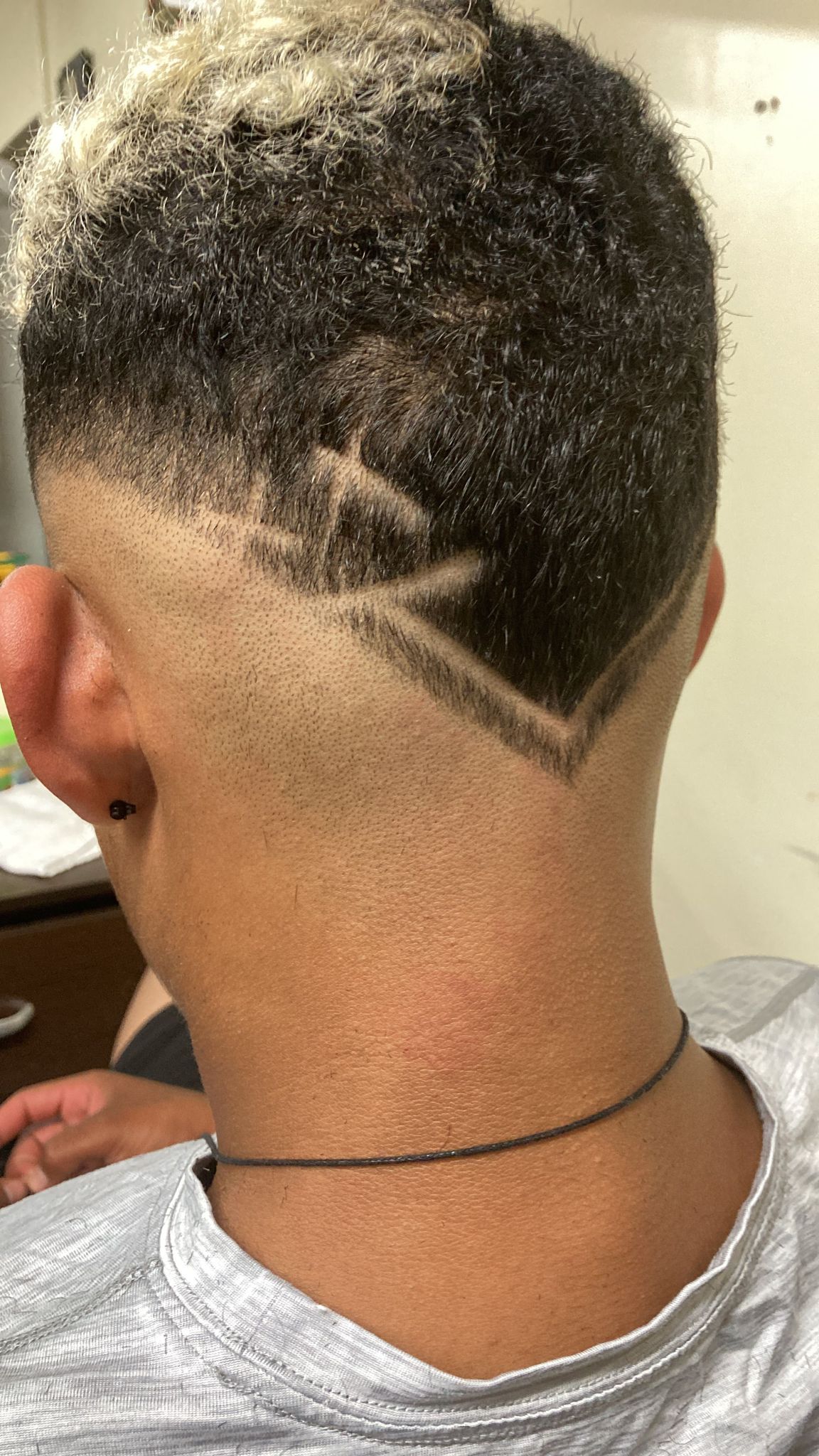 A young man has a haircut featuring a high fade with the top hair left longer and dyed another color. The sides and back are closely shaved with a geometric design, including sharp lines and angles shaved into the hair. The design starts from the middle of the back of the head and extends down towards the nape of the neck.