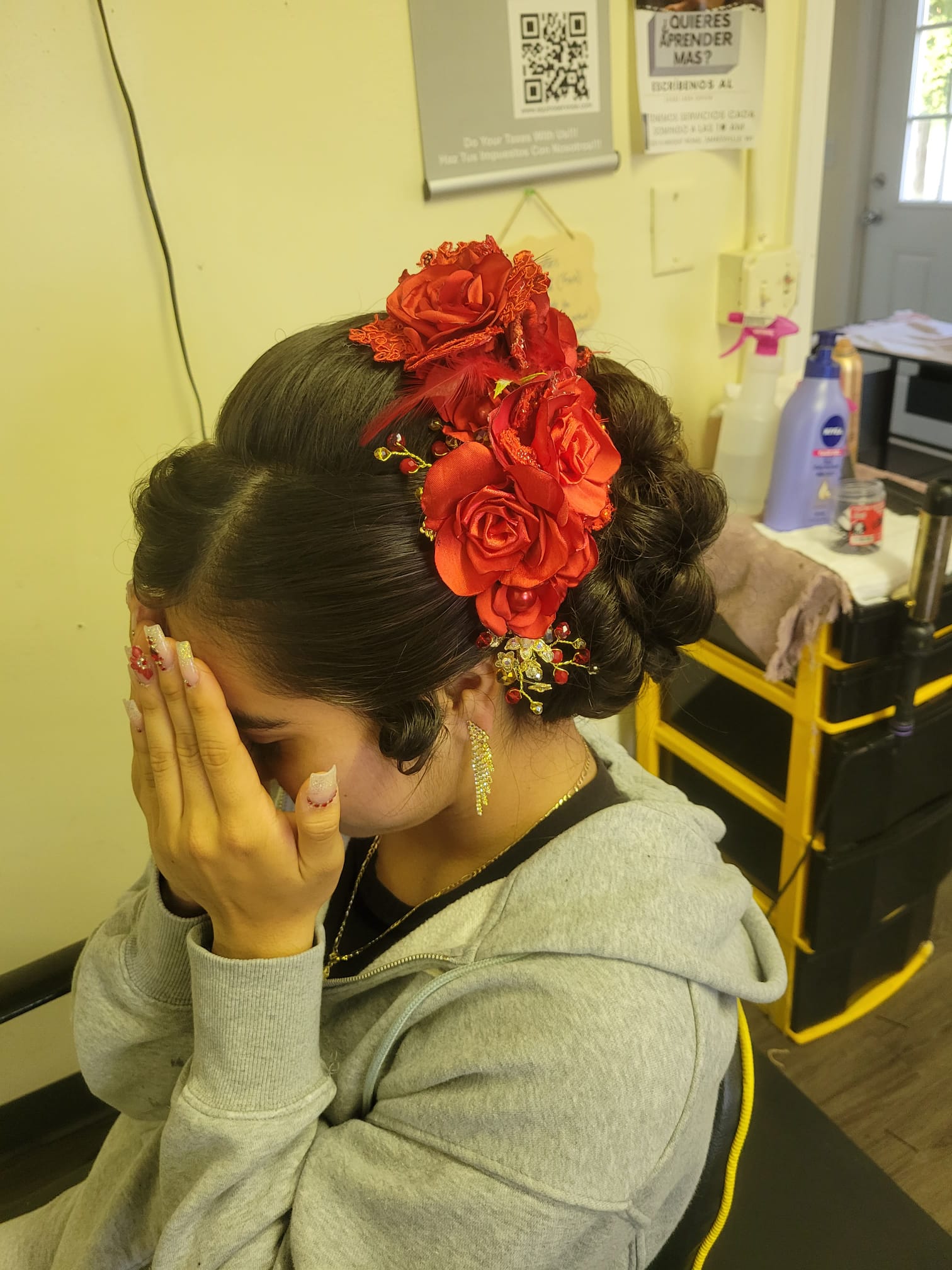 A young woman is wearing a Floral Braided Bun hairstyle. The hair is elegantly styled into a low bun with tight braids coiled around it. Large fabric flowers and decorative embellishments are pinned to the bun, adding a vibrant and festive touch. The woman is covering her face with her hands, showcasing her detailed nail art.