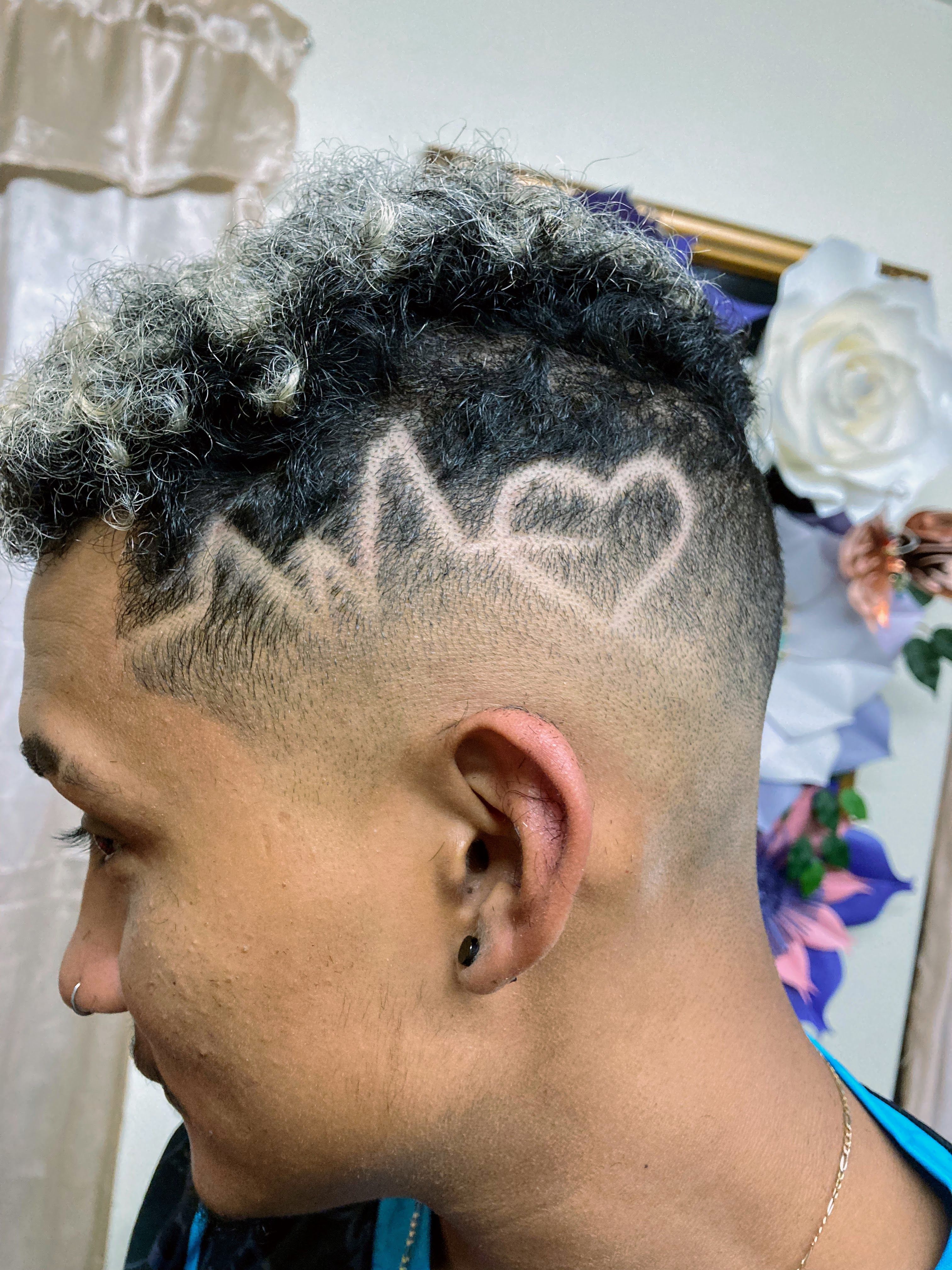 Man with a short haircut featuring a geometric design shaved into the side. The design includes a stylized zigzag pattern that strikes through a heart shape. The top of the hair is slightly longer and curly, while the sides are closely shaved for contrast.