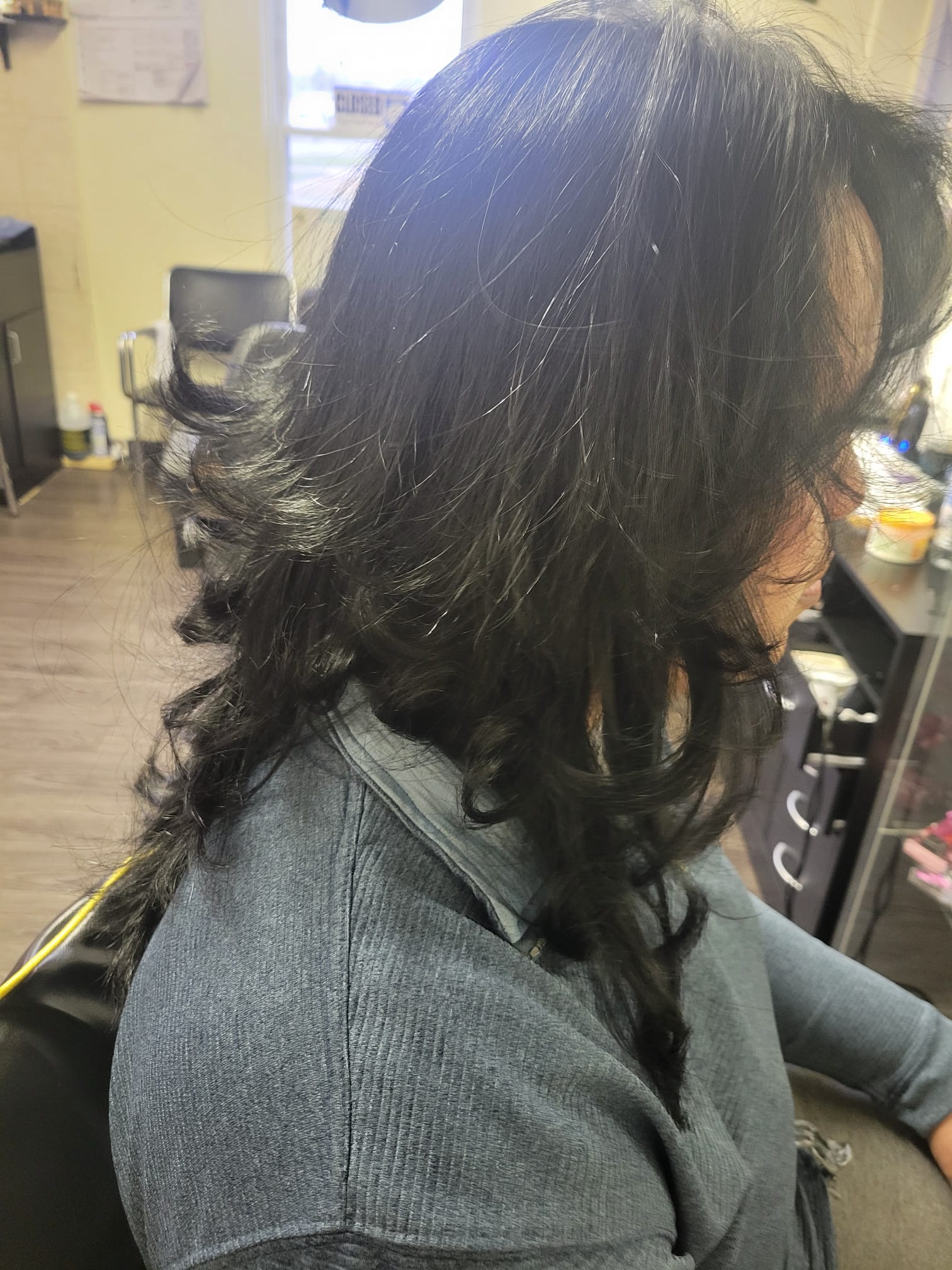 A side view of a person with long, layered hair styled with outward feathered ends, creating a voluminous and flowing look. The hair shines with a healthy gloss, and the layers give a textured, dynamic appearance.