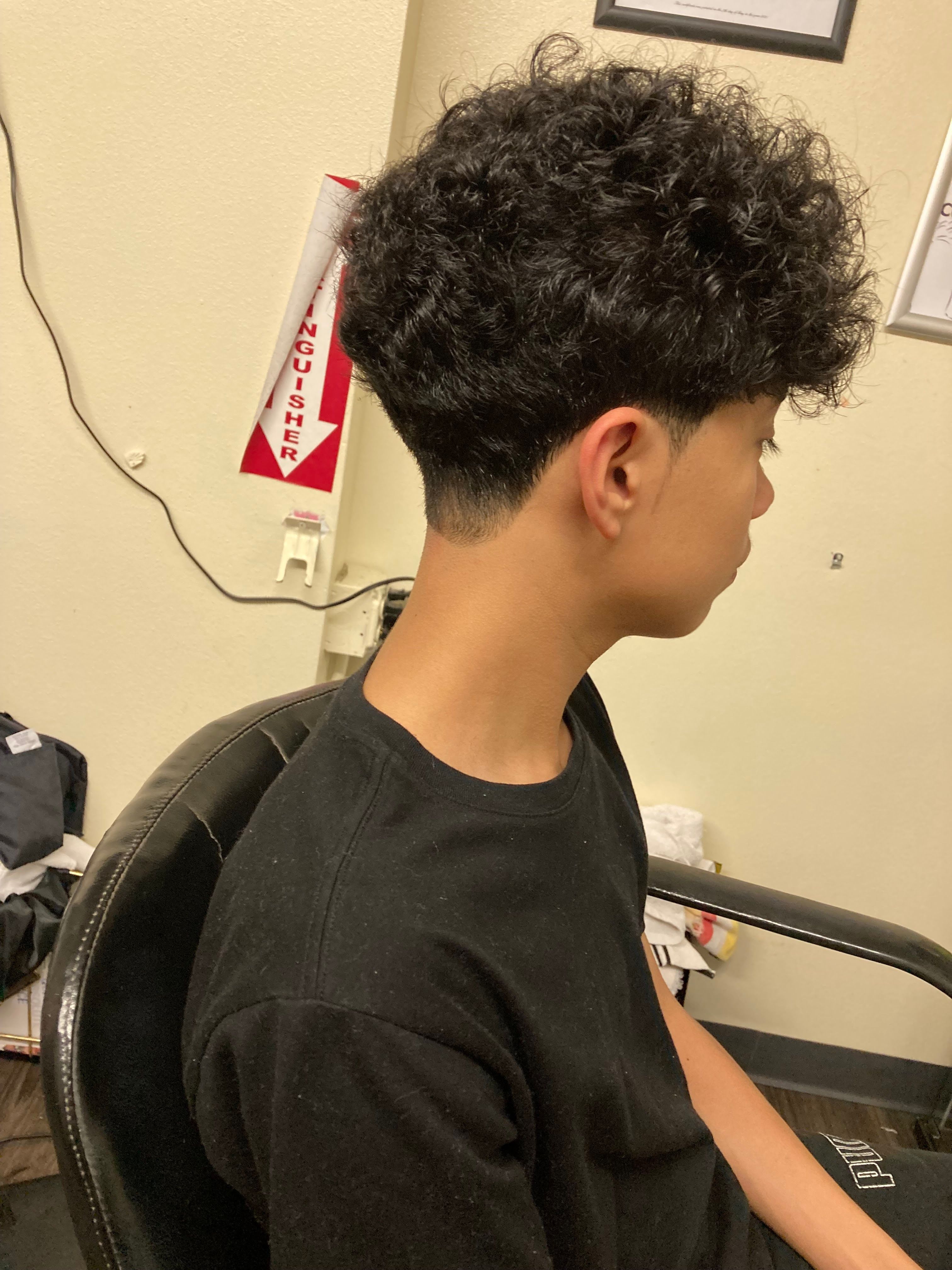 A young person with thick, naturally curly black hair styled in a low taper fade haircut. The curls are voluminous on top, while the sides and back are gradually tapered close to the skin at the nape, blending cleanly into the curls.