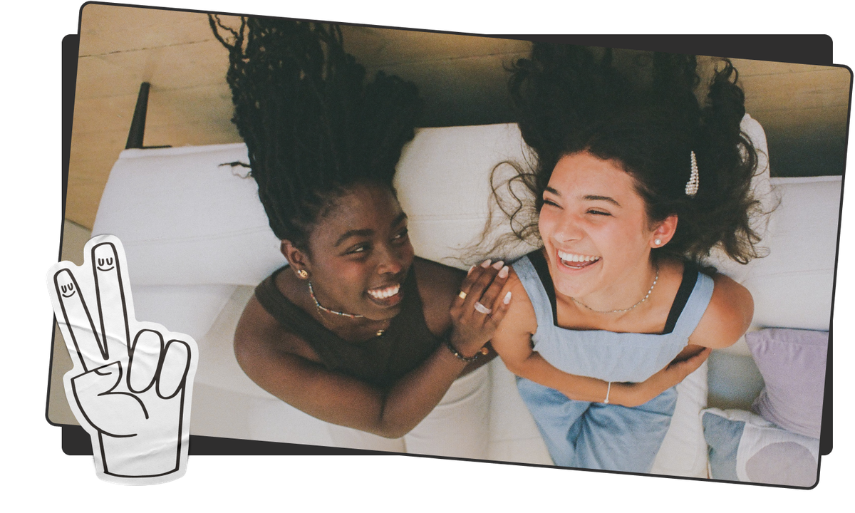 alt="Photo of two girls laying upside down on a couch and laughing together with a sticker of a peace sign on the left."