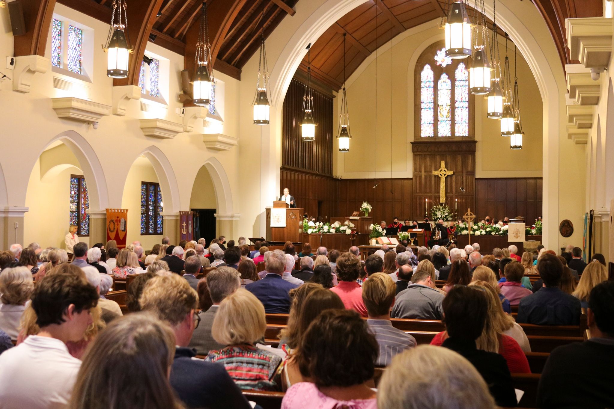 The SMCC sanctuary with white plaster walls, white stone side colonnades, and a wood panelled front apse and arched ceiling. A woman preaches from the dark wood pulpit at the front left of the apse. The pews are filled with people.