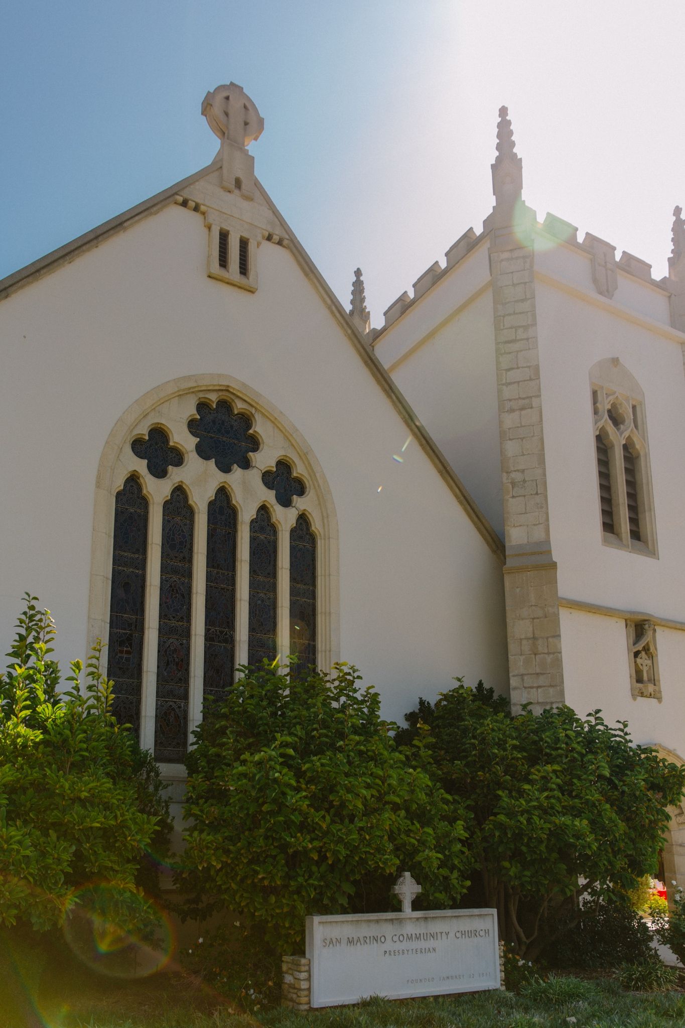 The west facade of SMCC. Five narrow stained glass windows are crowed by three rose windows under a pointed roof and a stone cross, next to the square bell tower.