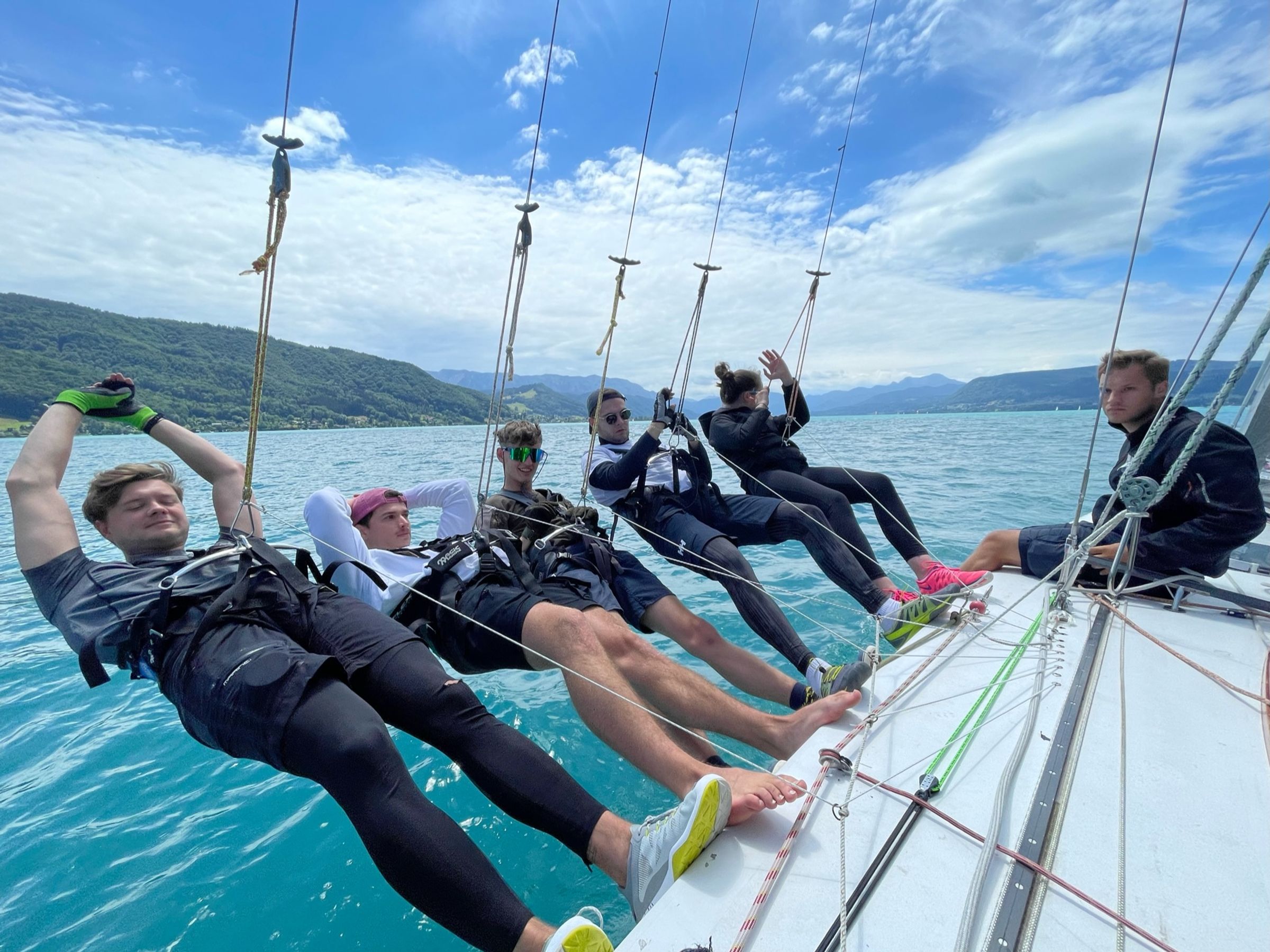 Die Crew im Trapez weit über der Bordkante, dahinter der türkise See und die Berge am Ufer