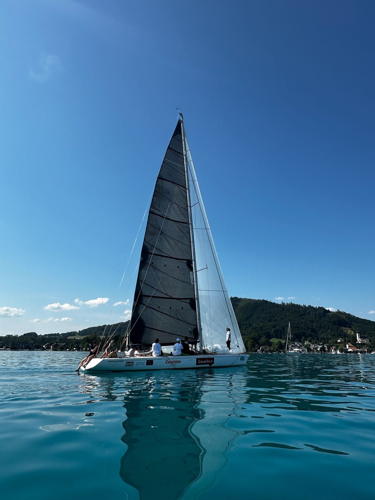 Liberté in light airs off the village of Attersee, her dark mainsail against a blue sky