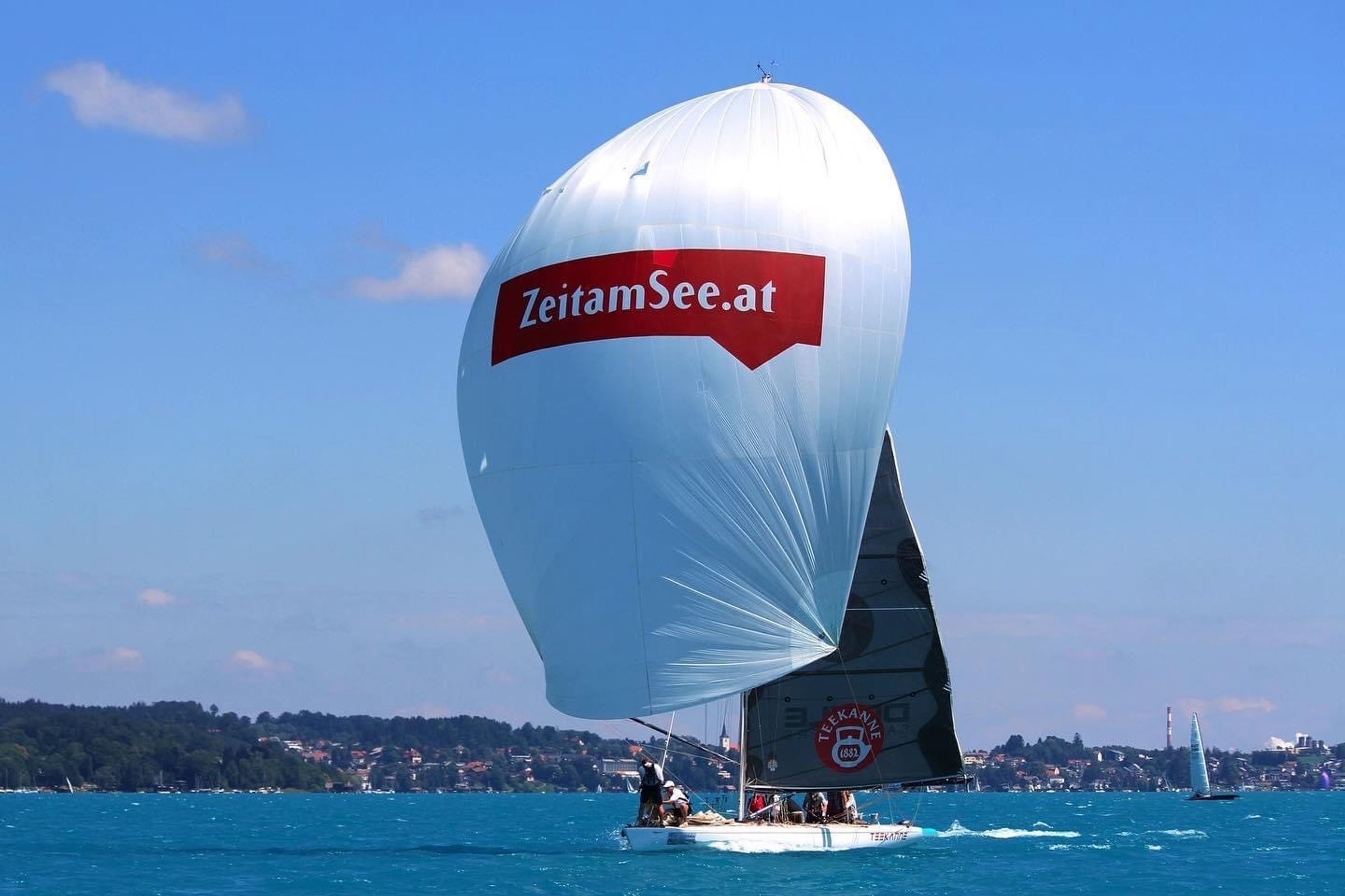 Der weiße Spinnaker mit dem Schriftzug ZeitamSee.at unter blauem Himmel, im Großsegel das Logo von Teekanne