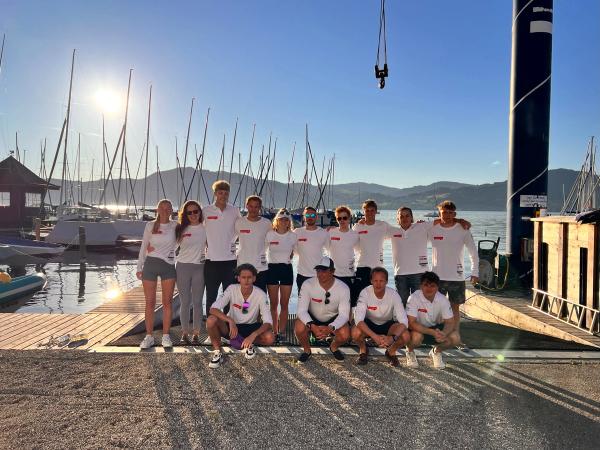 Vierzehn Seglerinnen und Segler in weißen Teamshirts am Hafen im Morgenlicht, dahinter Masten und der See