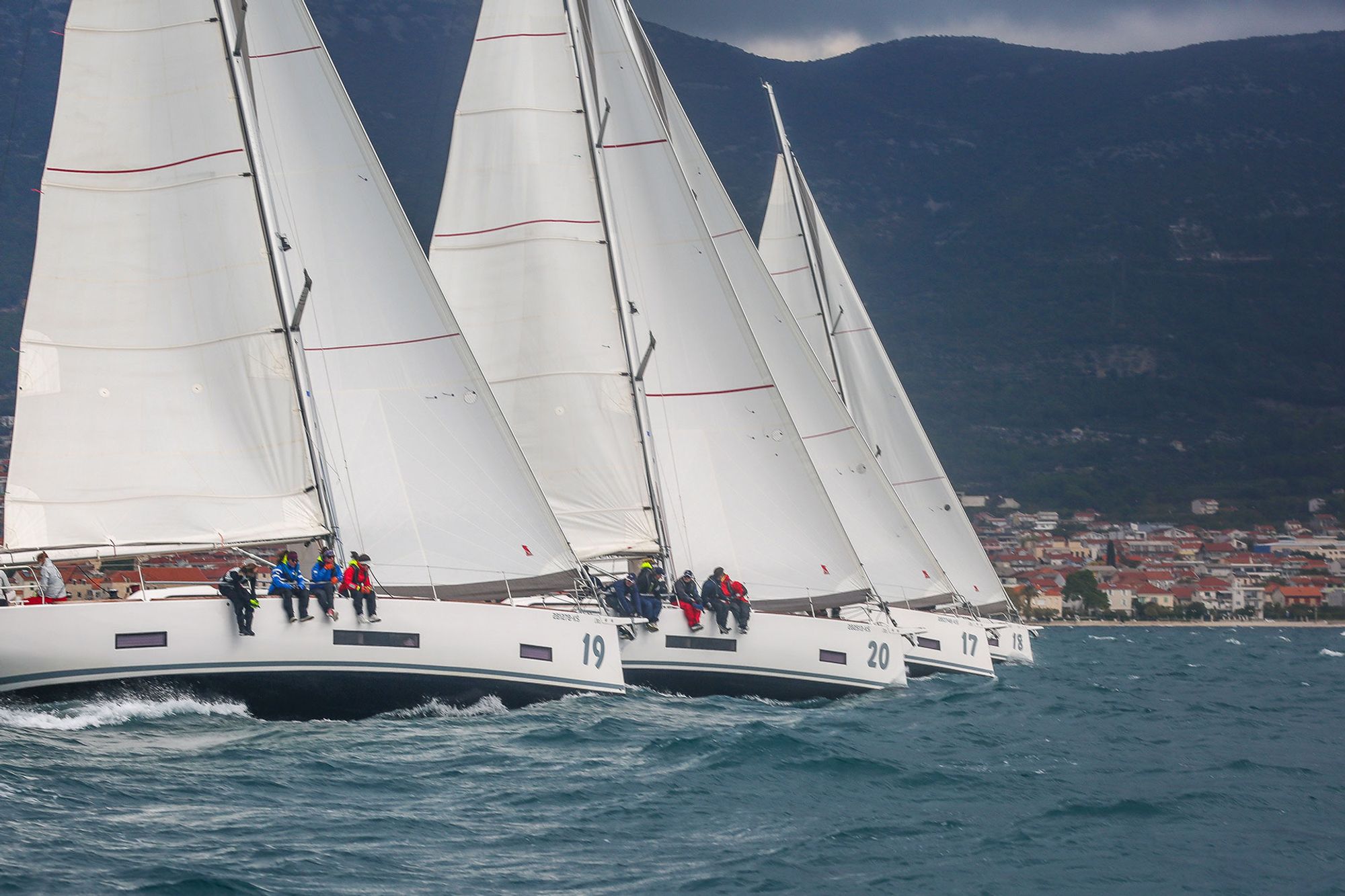 Vier baugleiche Yachten kurz nach dem Start in einer Linie, dahinter die Küstenberge