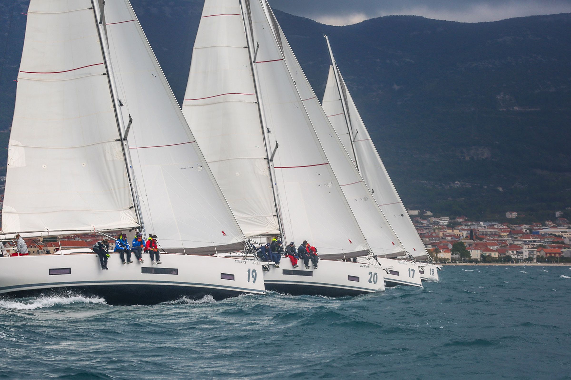 Four identical yachts in line shortly after the start, coastal mountains behind