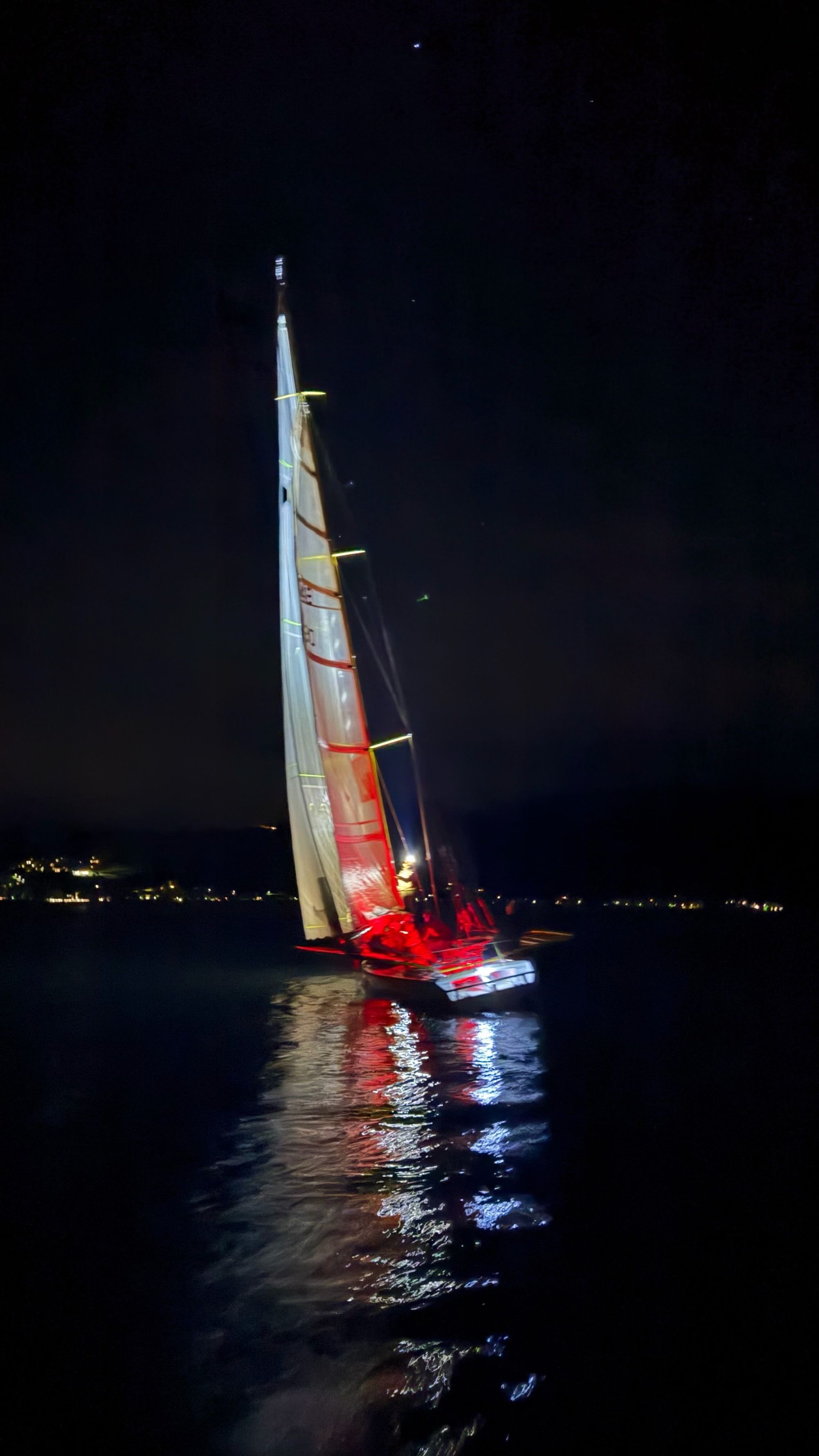 Ein Boot unter Segeln bei Nacht, die Lichter spiegeln sich im Wasser