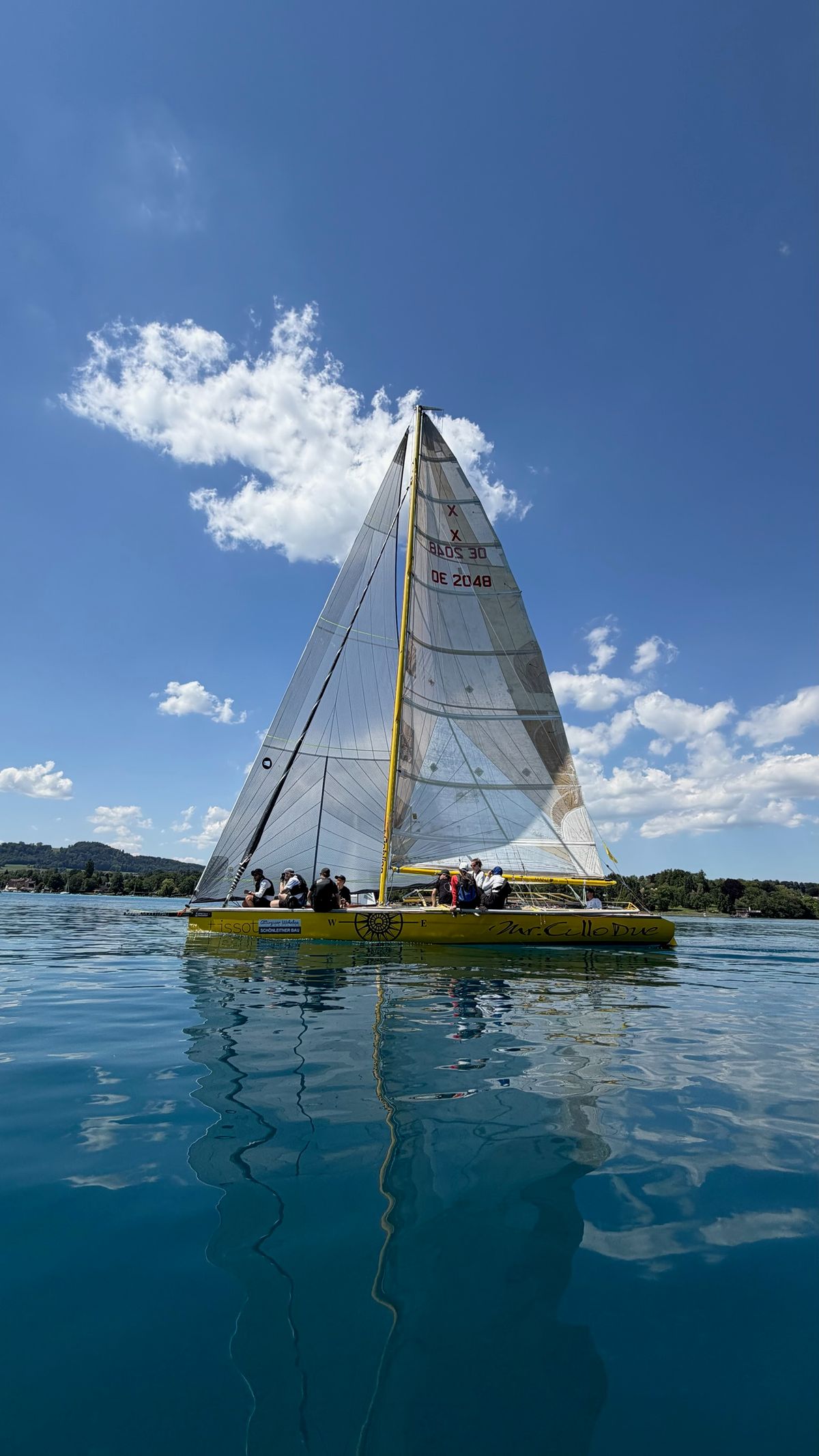 Mr. Cello Due becalmed on Lake Attersee, sail number OE 2048 on the headsail, her reflection in the water