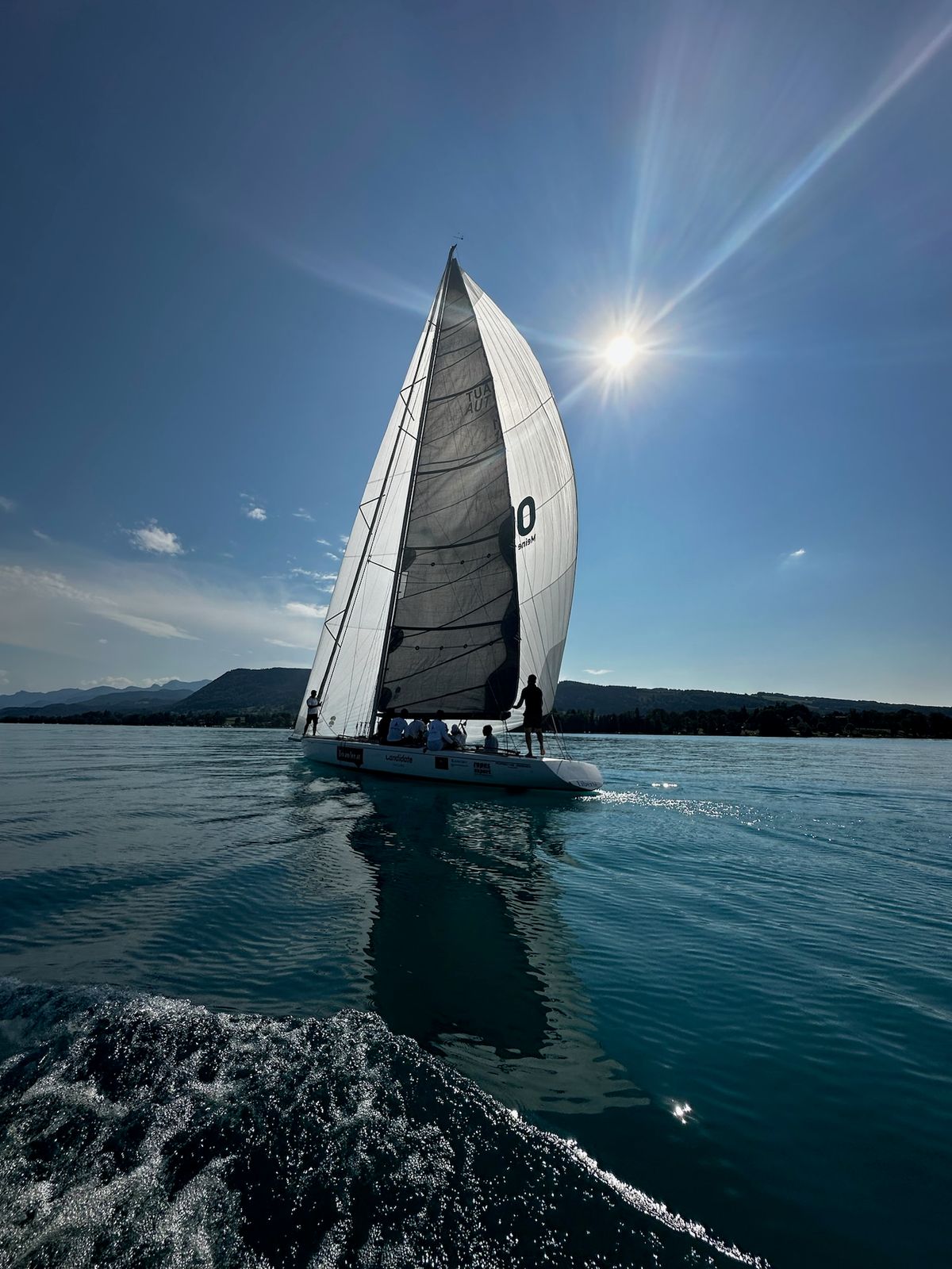 Liberté under full sail, backlit by the sun on Lake Attersee, crew on deck
