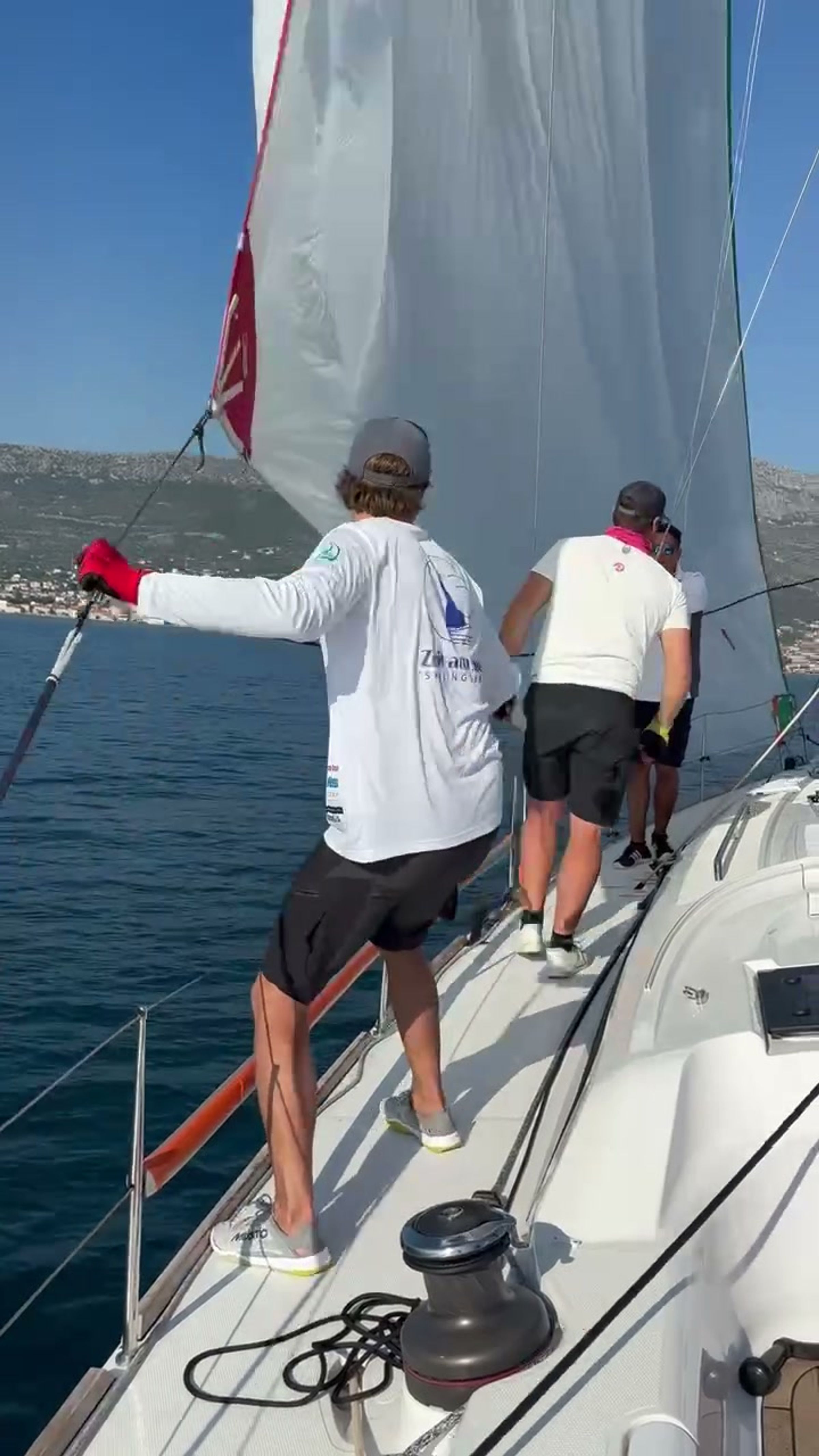 Crewmitglied mit dem Schriftzug Zeit am See Sailingteam auf dem Weg nach vorn, dahinter die Küste von Kaštela