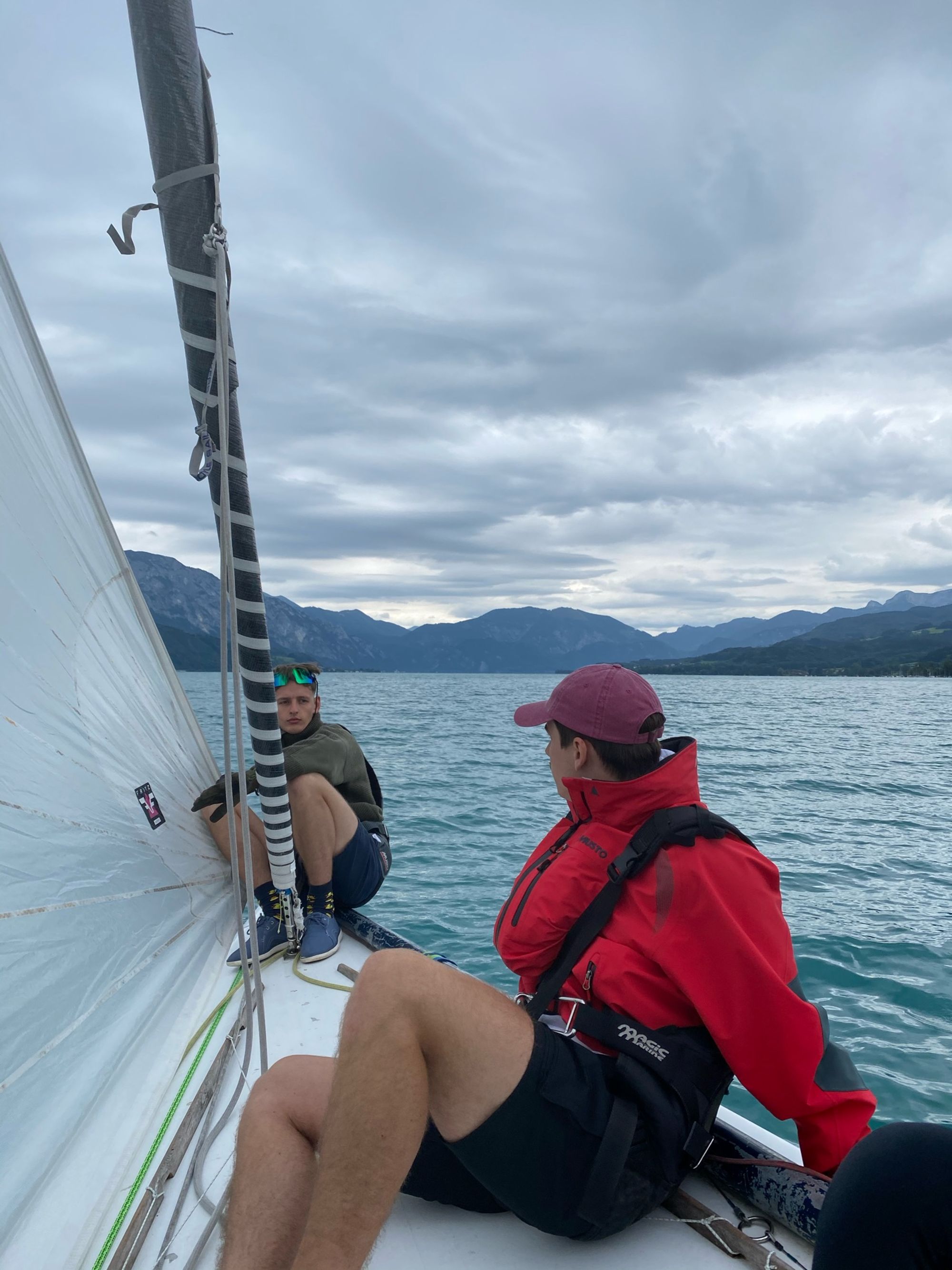 Zwei aus der Crew auf dem Vorschiff, einer in roter Jacke, dahinter der See und die Berge unter grauem Himmel