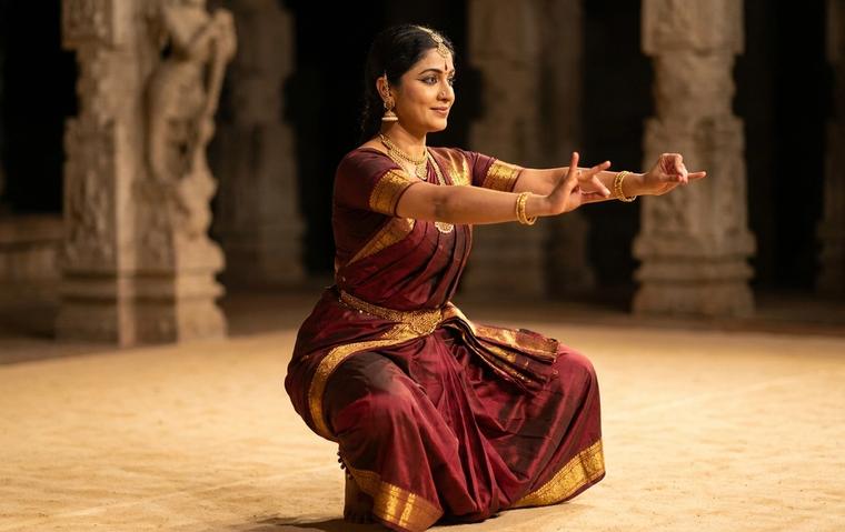Bharatanatyam dancer holding a deep aramandi stance in a maroon and gold costume, showing the clean lines that distinguish one bani from another.
