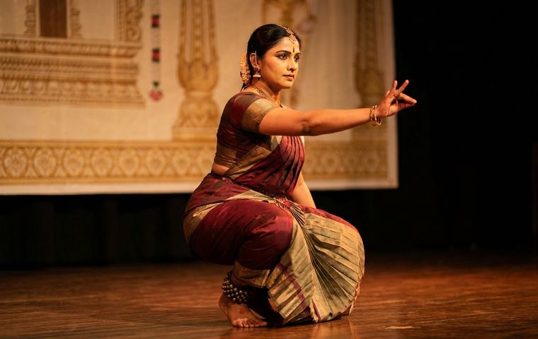 Bharatanatyam dancer in deep aramandi performing a varnam on a warm-lit stage in maroon and gold costume