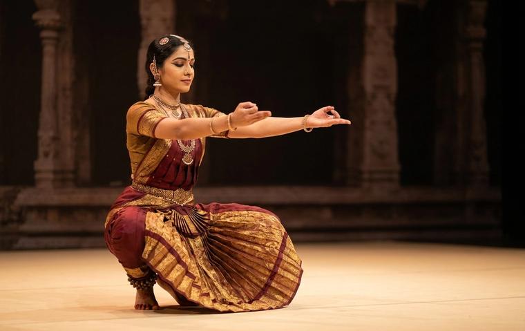 Bharatanatyam dancer holding a deep aramandi posture in maroon and gold costume on a warm lit stage