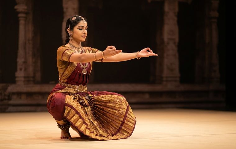 Bharatanatyam dancer holding a deep aramandi posture in maroon and gold costume on a warm lit stage