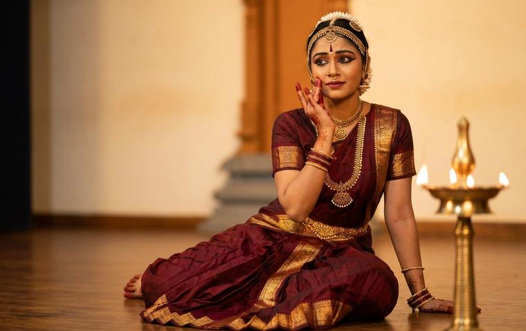 Bharatanatyam dancer in maroon and gold costume performing abhinaya during a padam, her expression and hand gesture caught in warm lamp light